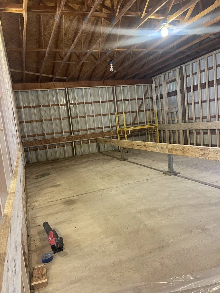 Interior of an unfinished garage with exposed wooden framing, spray foam insulation, a concrete floor, and a partially built wooden wall. A yellow scaffold is in the corner, and a cordless drill and tape measure sit on the floor.