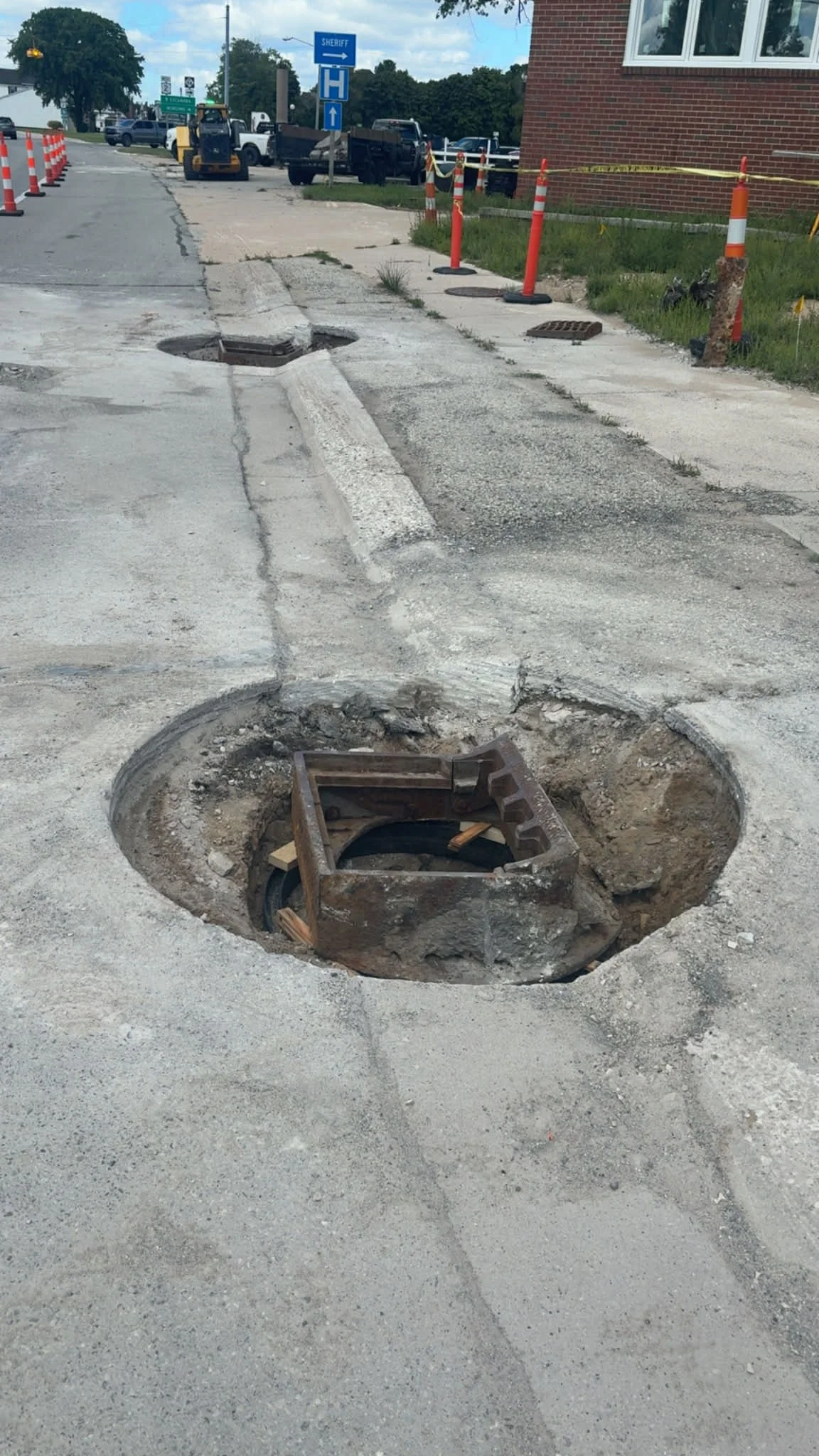 Road construction site with open manholes, orange cones, and construction equipment in the background.