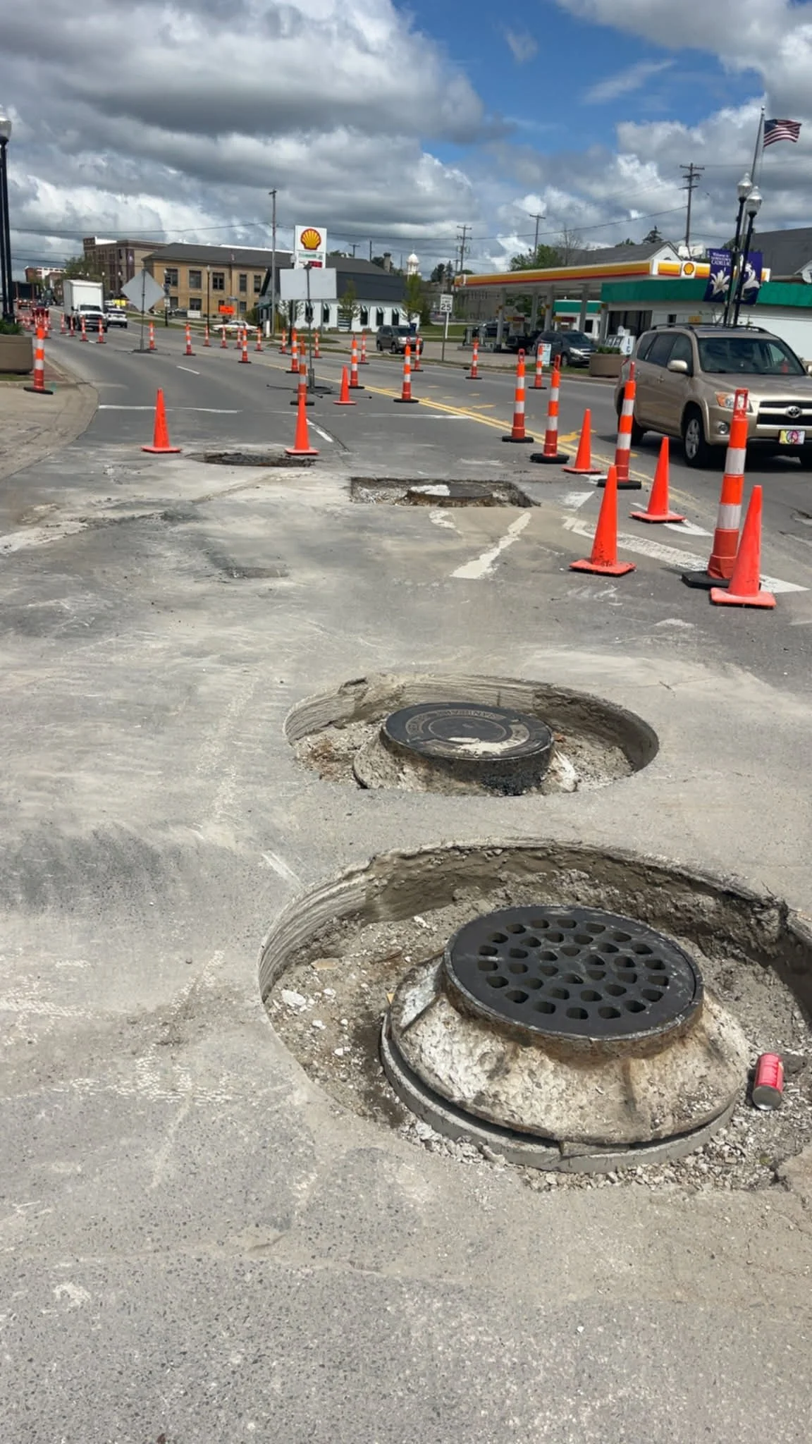 Street with construction cones and open manhole covers, traffic and a Shell gas station in the background on a partly cloudy day.