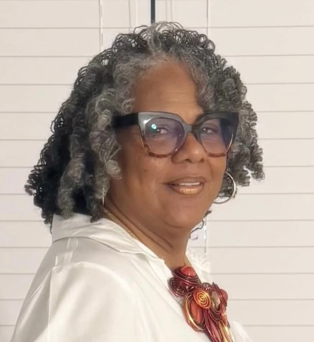 A woman with curly gray hair, wearing large glasses, gold earrings, a white outfit, and a red necklace, standing against a white horizontal paneled background.