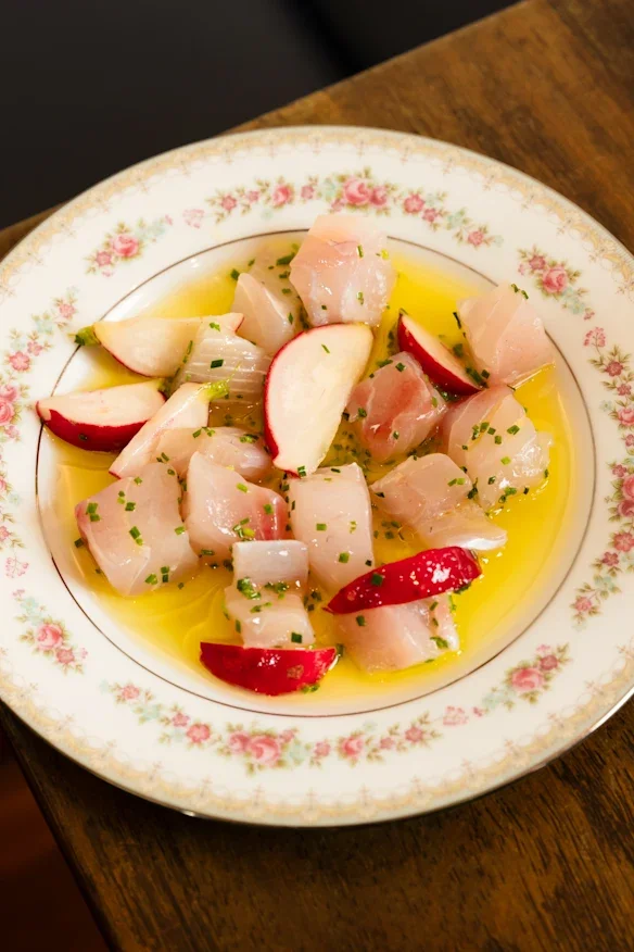 A decorative floral-patterned plate with cubed raw fish, apple slices, and chopped chives, garnished with olive oil.