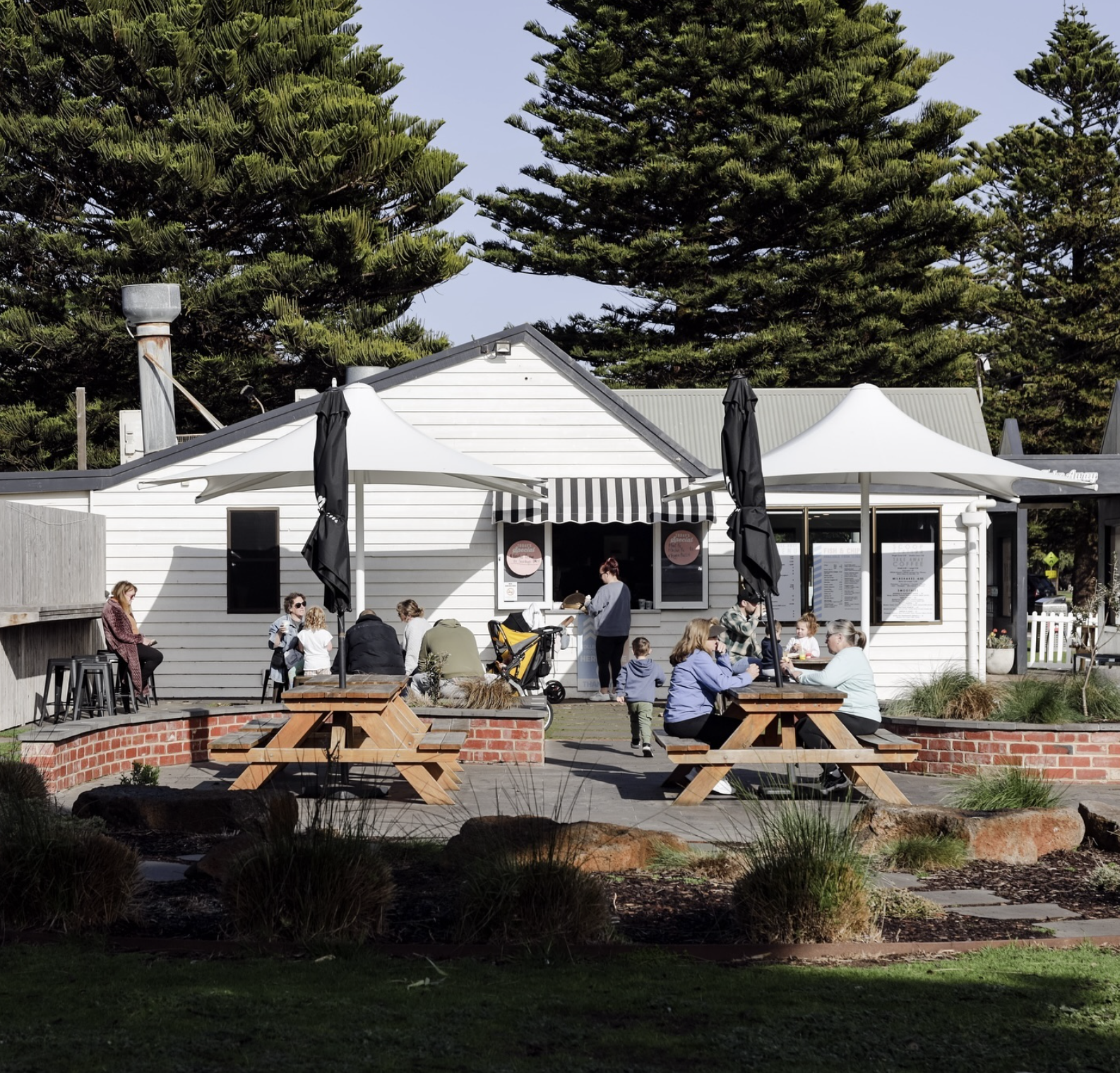 A group of people sitting and socializing outside a small white building with striped awnings and umbrellas, surrounded by greenery and tall trees.