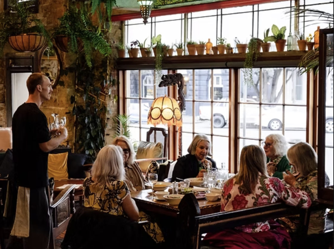 A group of six elderly women having a meal together in a cozy restaurant with large windows, plants, and decorative lighting, while a man stands nearby holding a glass.