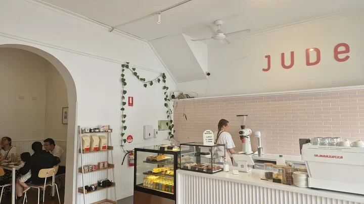 Interior of a cafe named Jude, with a display case of baked goods, a barista at the coffee machine, and customers seated at tables.