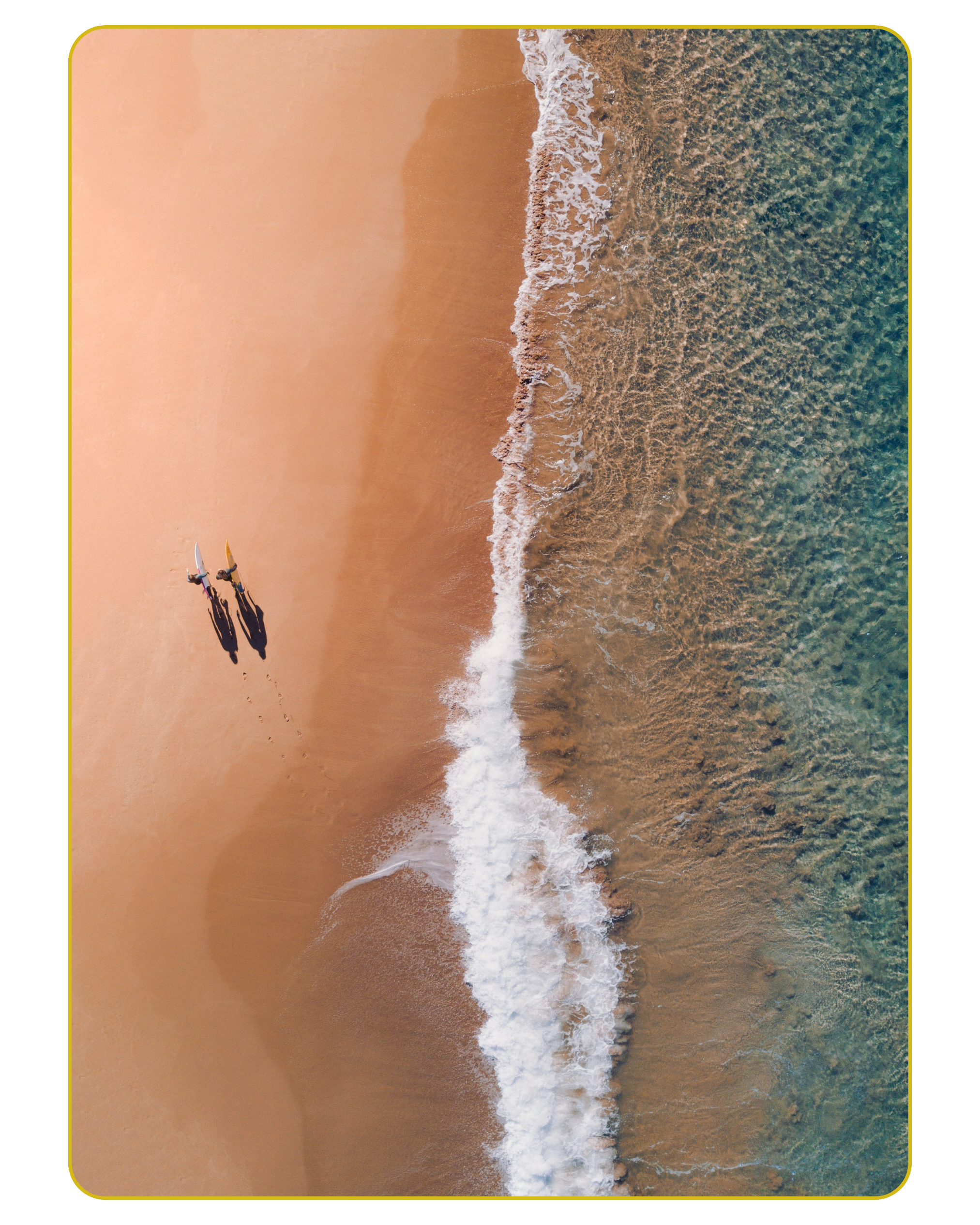 Bird's eye view of a sandy beach with gentle waves and three surfers walking toward the water, casting long shadows.