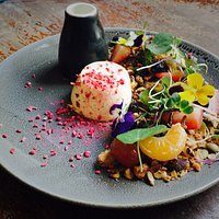 Dessert plate with a scoop of pink and white ice cream, fresh fruits, edible flowers, nuts, and a drizzle of sauce on a gray plate.