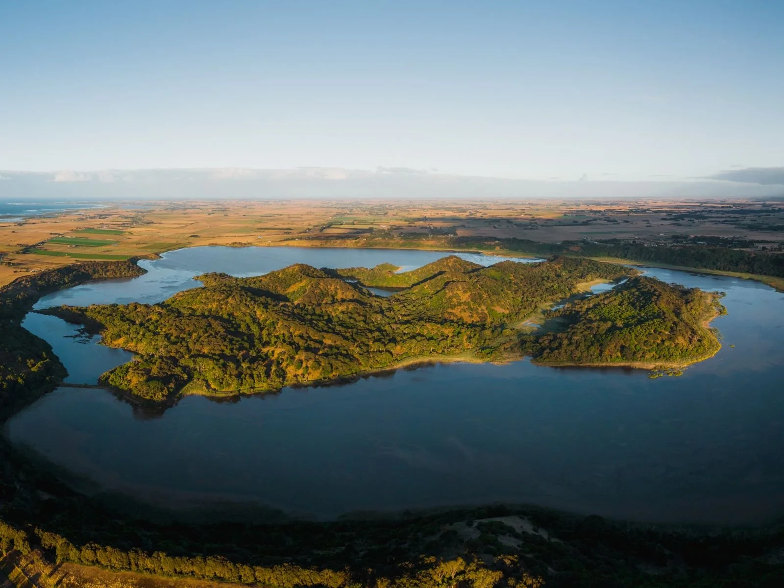Aerial view of a large island surrounded by water with lush green forests and hilly terrain, with open farmland in the background under a clear blue sky.