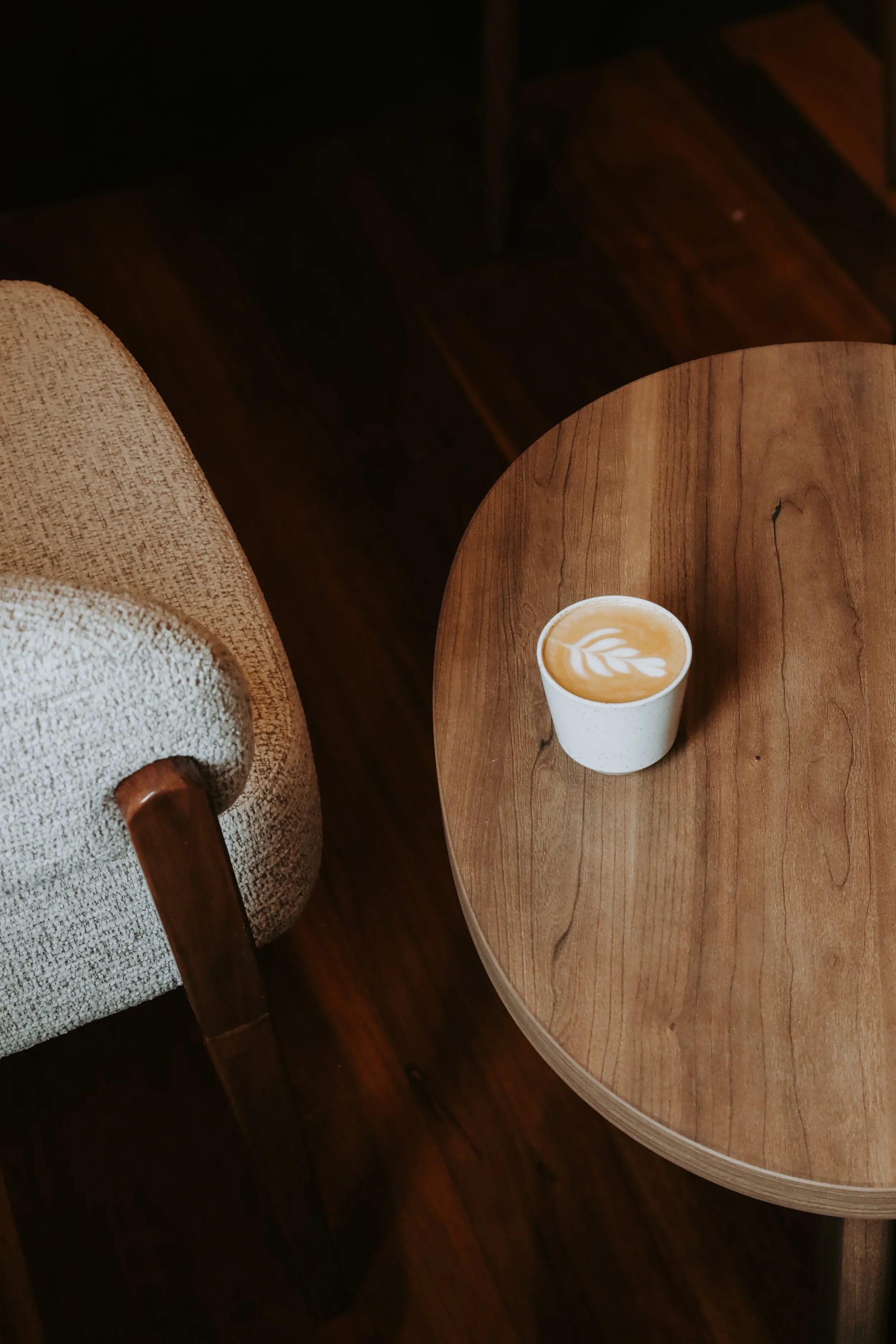 A latte coffee with latte art on top sitting on a round wooden table in a cozy cafe. Part of a beige upholstered chair is visible on the left side of the image.