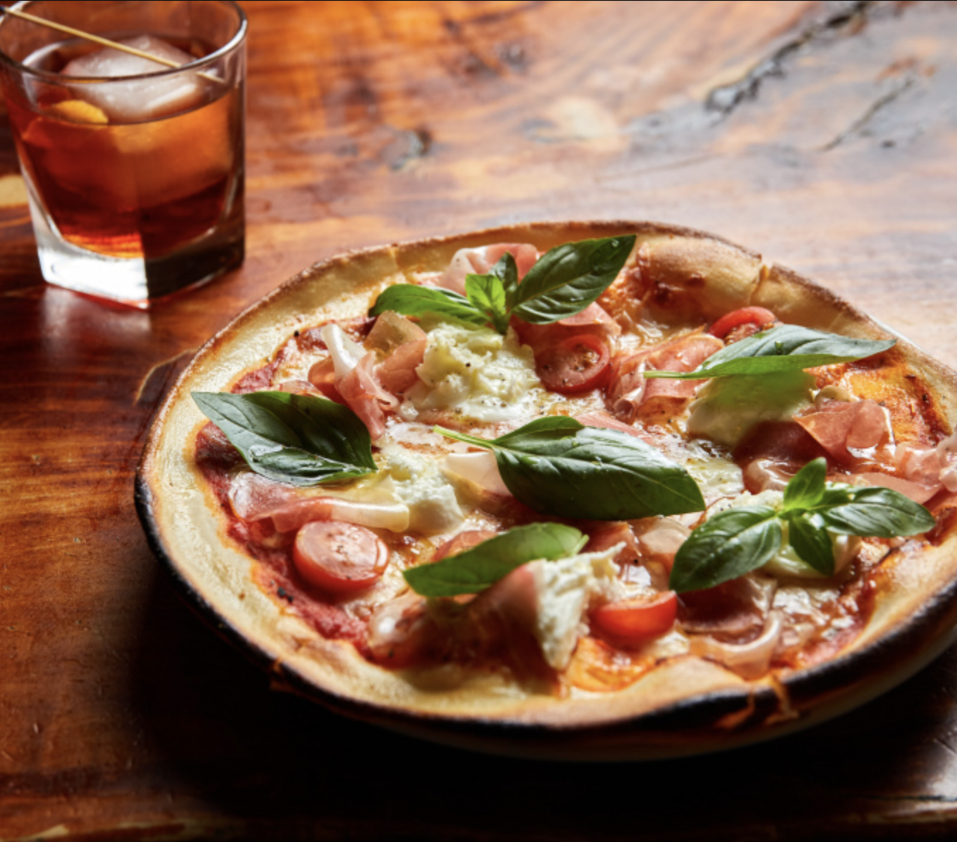 A pizza topped with fresh basil leaves, cherry tomatoes, mozzarella cheese, and prosciutto, with a glass of dark soda with ice on a wooden table.