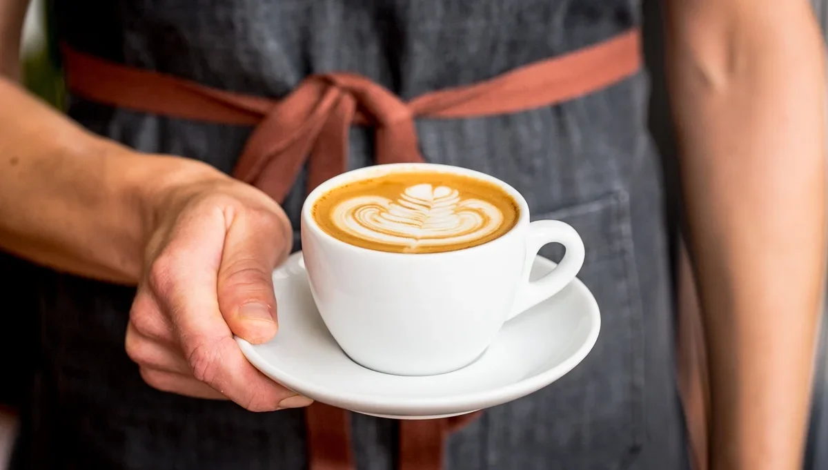 Person holding a white cup of coffee with latte art on top, wearing a gray apron tied with a brown strap.