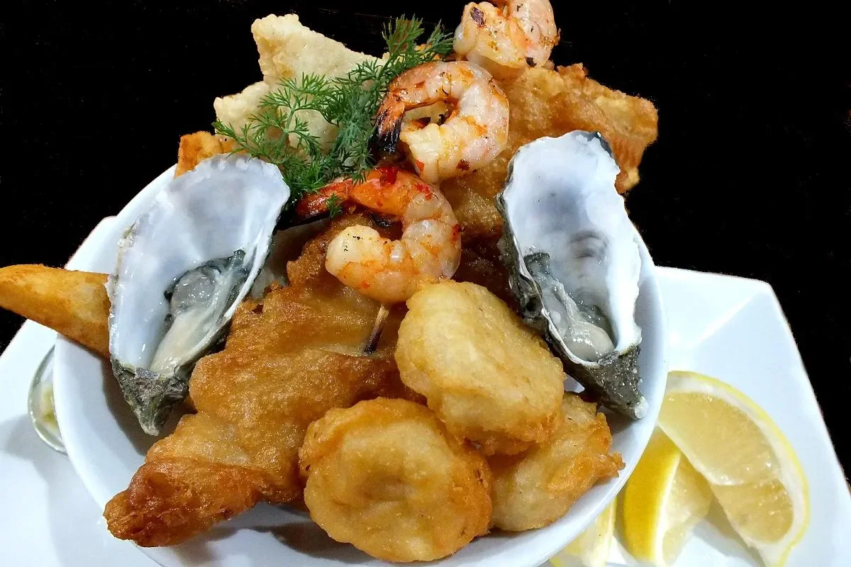Seafood platter with fried fish, shrimp, oysters, potato wedges, and lemon wedges.