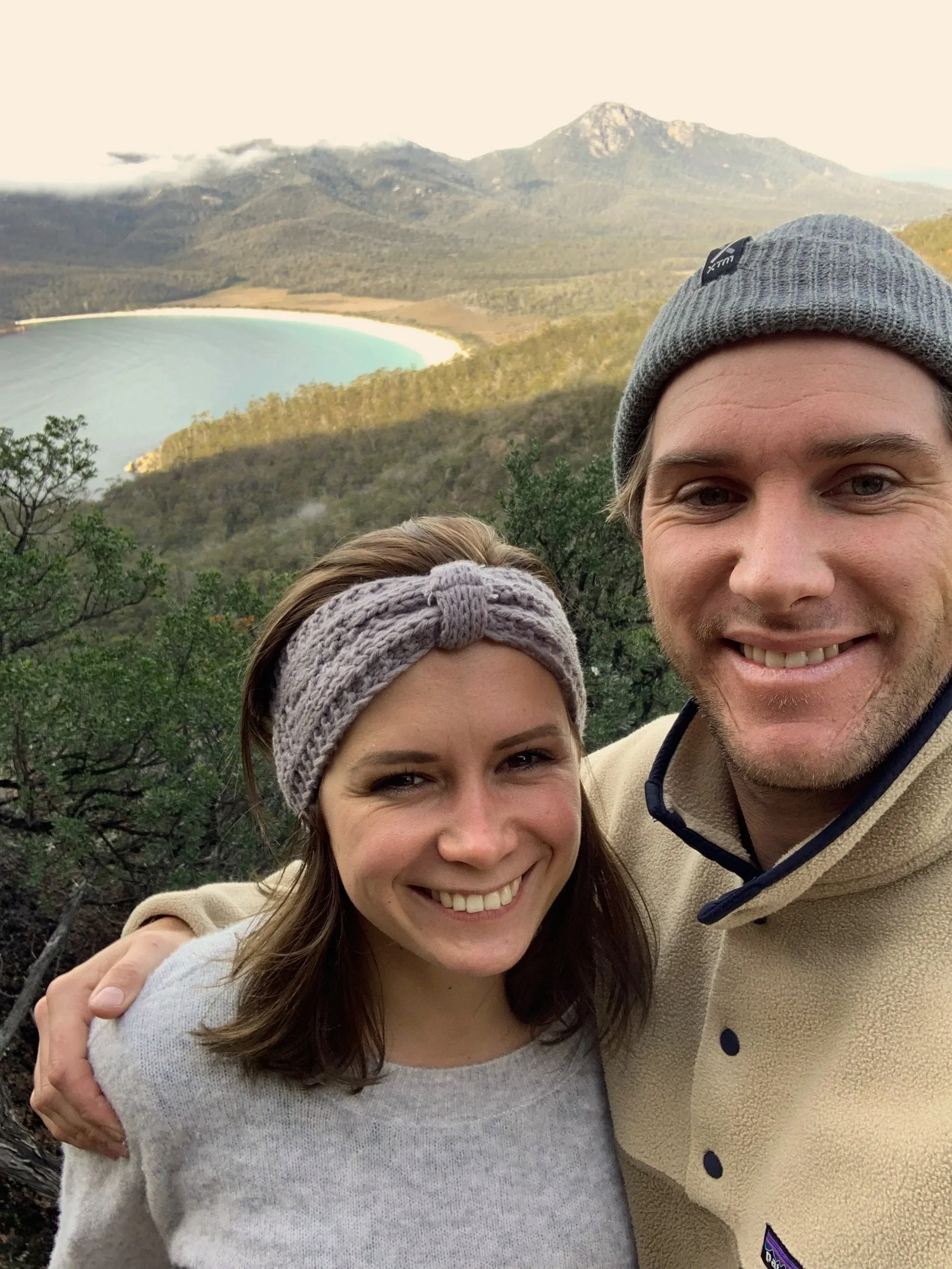 A smiling man and woman taking a selfie outdoors with a mountain, forest, and a body of water in the background.