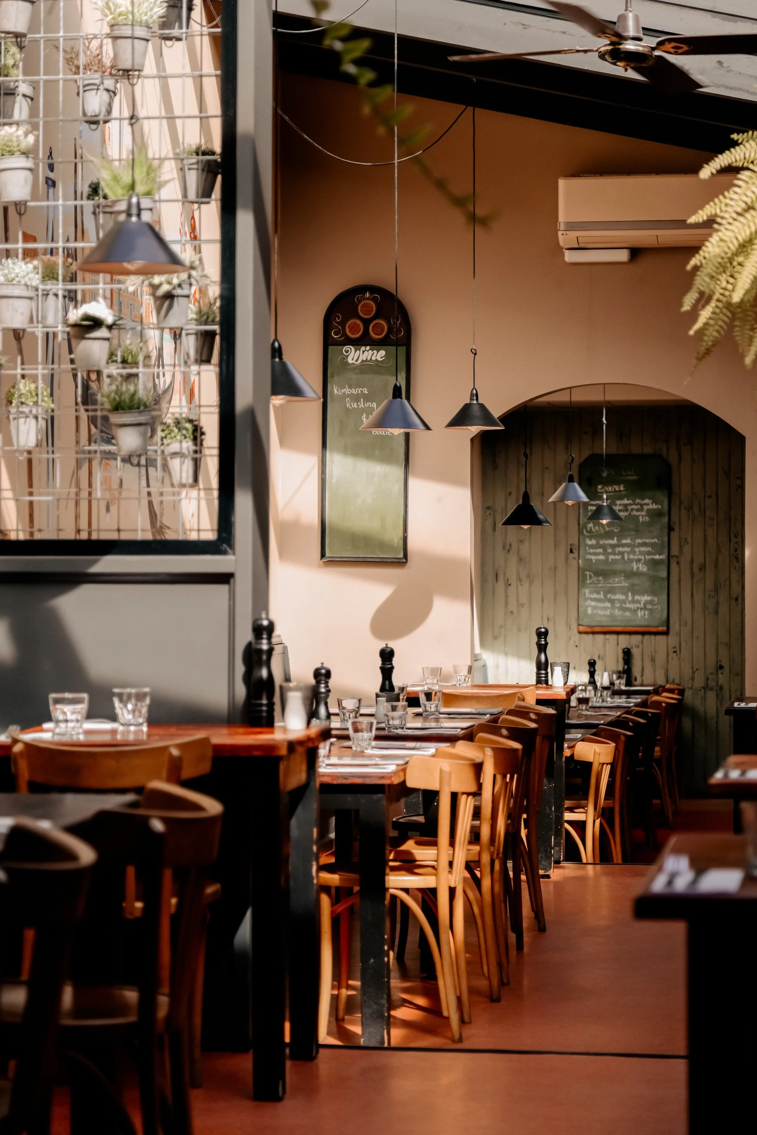 Interior of a cozy restaurant with wooden chairs, tables set with glasses and utensils, hanging black pendant lights, wall menu boards, and a relaxed ambiance.