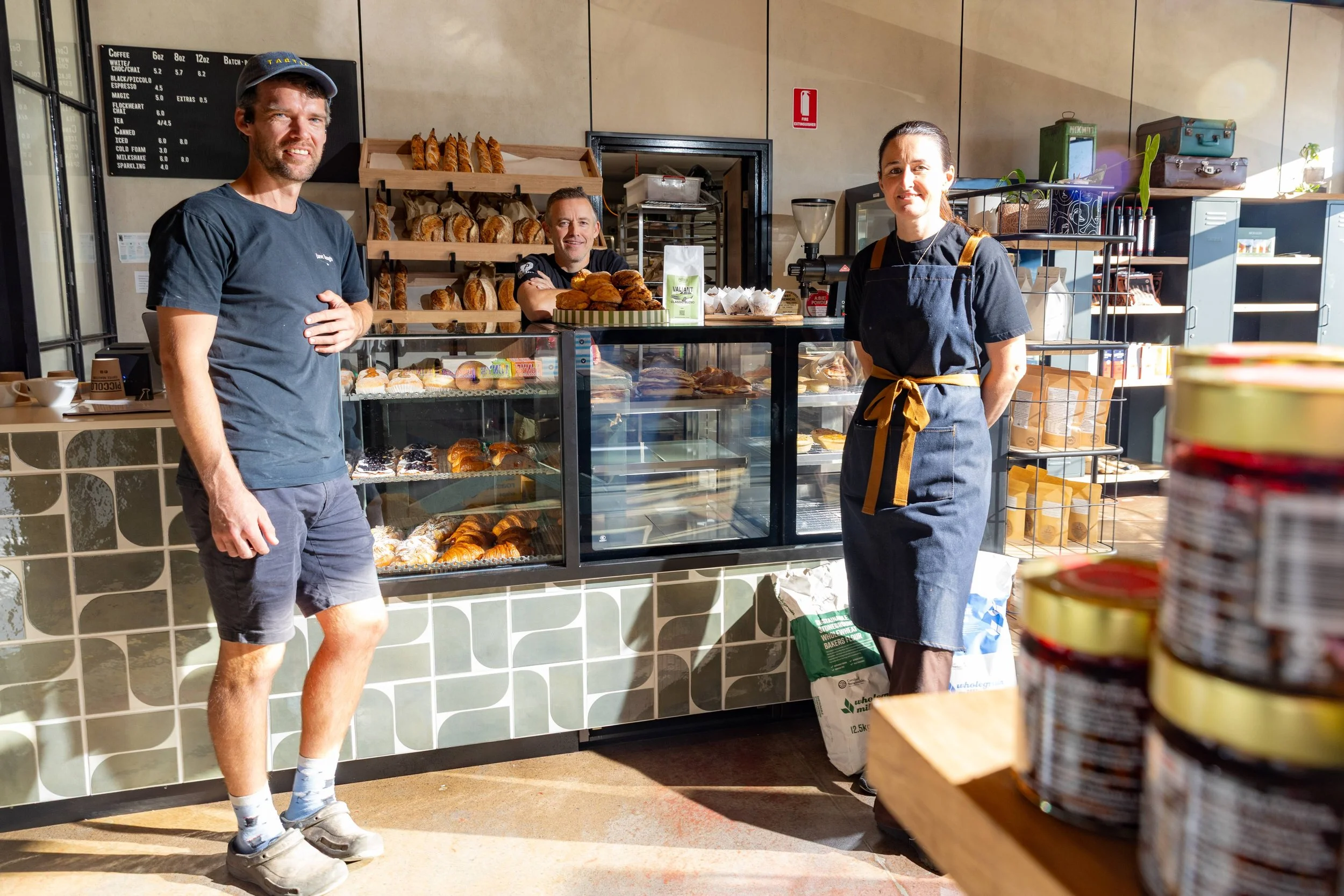 Three people standing inside a bakery or cafe, smiling at the camera. Behind them, there is a glass display case filled with various baked goods like croissants and pastries. Shelves with bread and other items are visible in the background, along with a menu board and some kitchen equipment. The person on the right is wearing a black apron with a brown tie, while the man on the left is casually dressed in shorts, a T-shirt, and a cap.