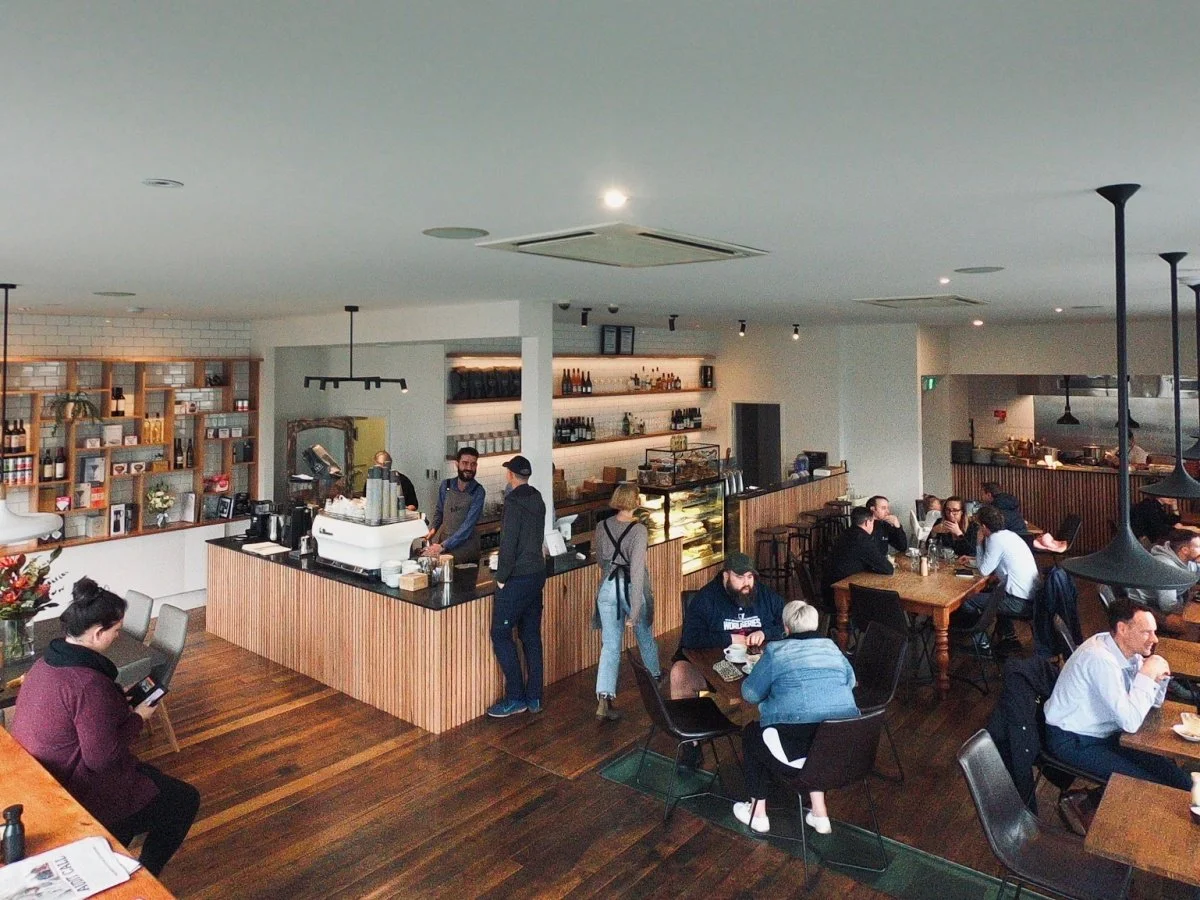 A busy cafe with customers sitting at tables and ordering coffee at the counter. Baristas are working behind the counter, and shelves with bottles and decor are visible on the wall.