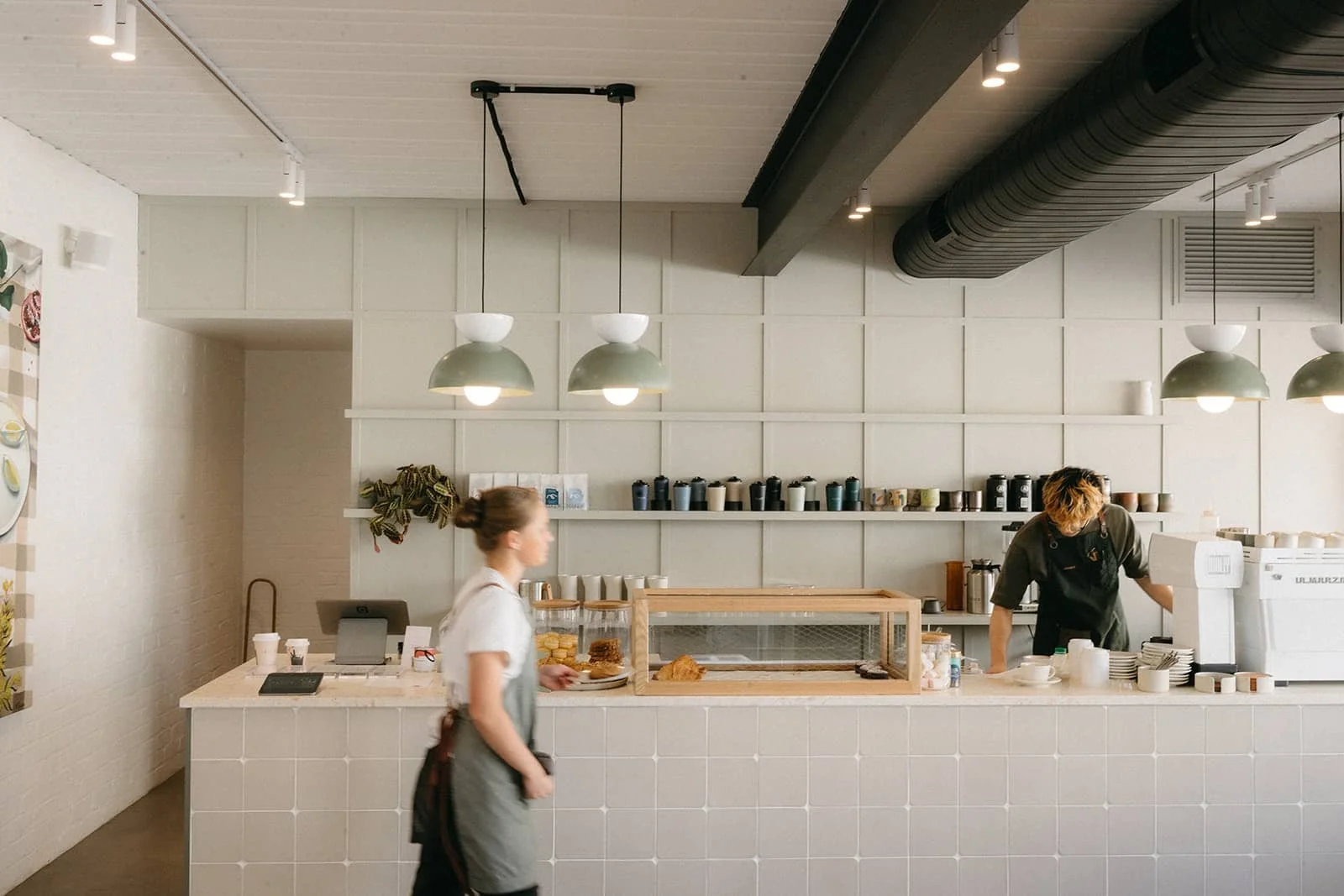 Interior of a modern cafe with a barista working behind the counter and a female customer at the counter. The cafe has minimalist decor with pendant lights, shelves with cups and containers, and a white tiled counter.