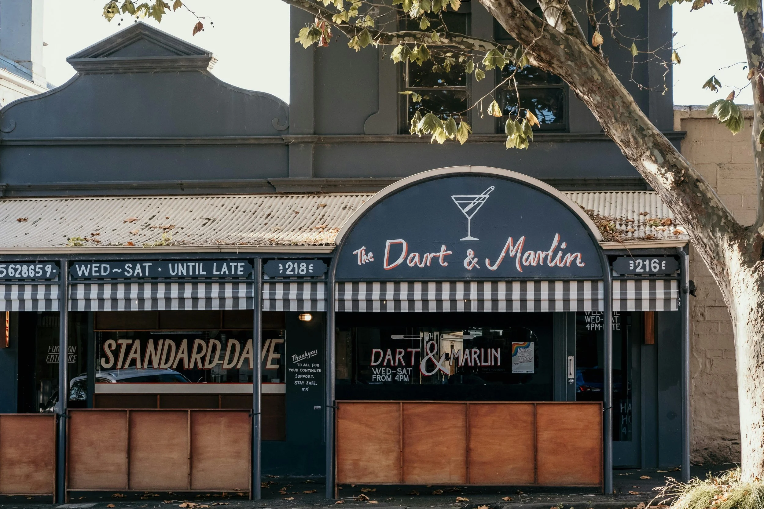 Storefront with a sign reading 'The Dart & Marlin' and striped awning, surrounded by a tree with green and brown leaves.