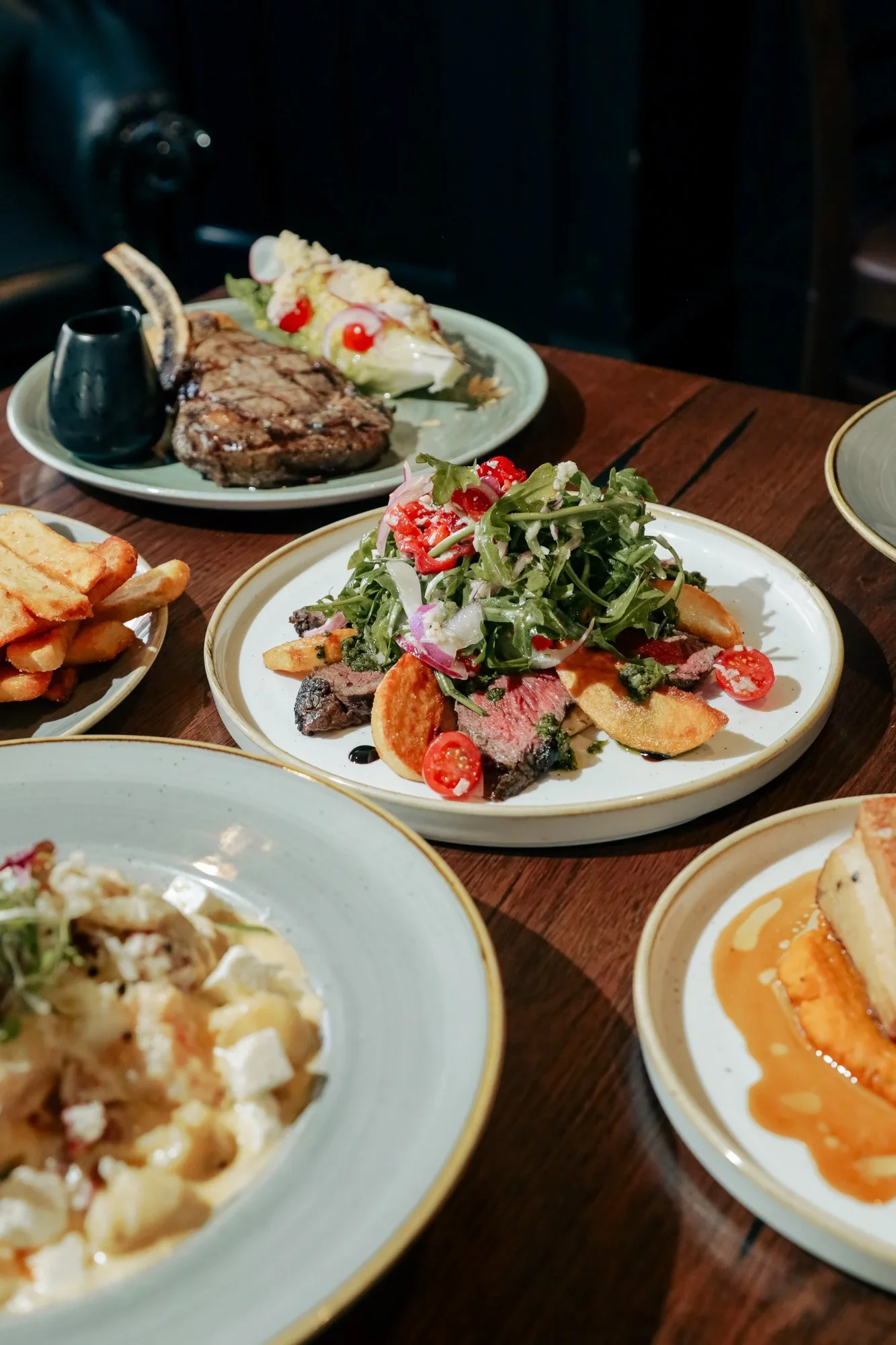Assorted dishes of food on a wooden table, including salad, steak, fries, and other items.