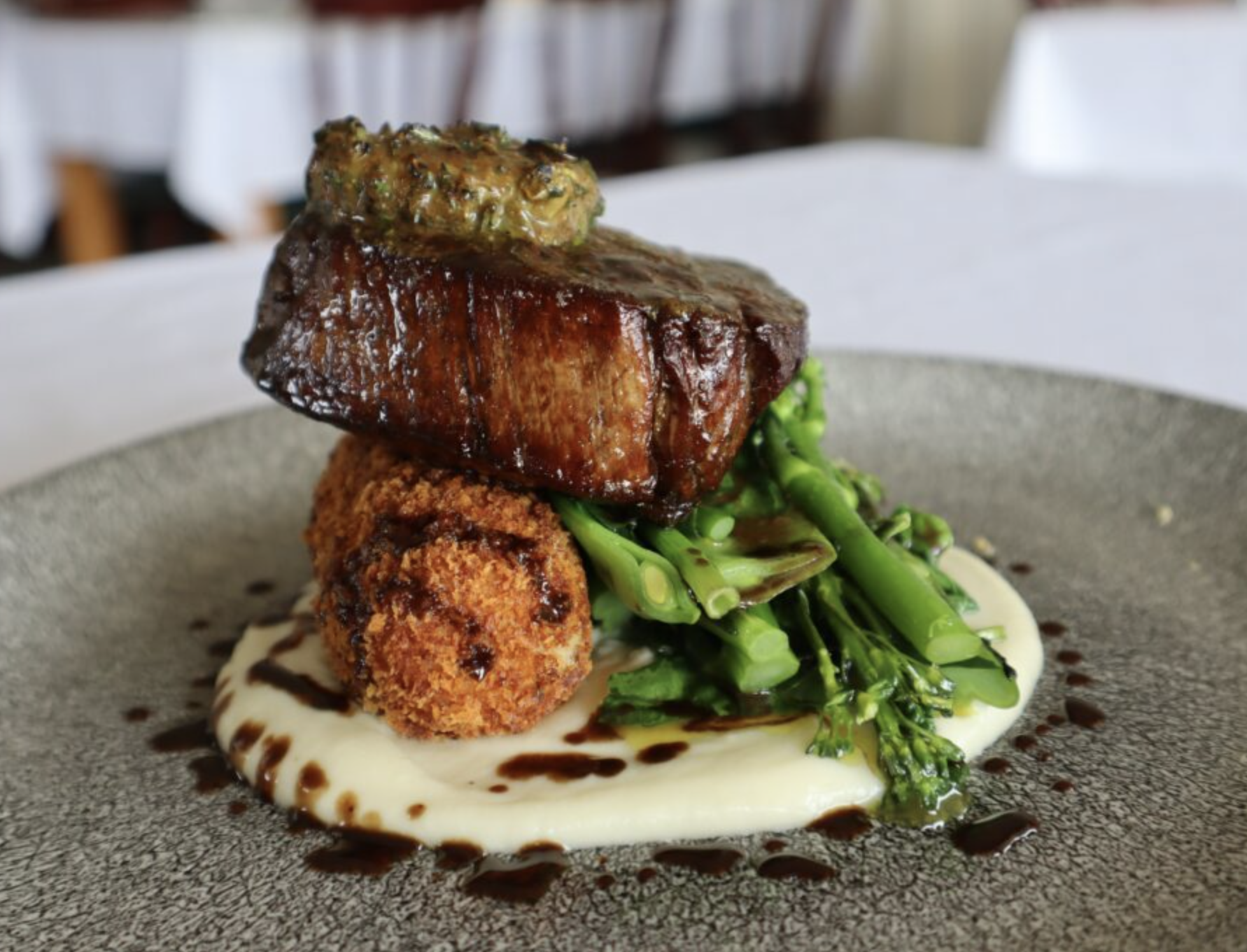 A gourmet dish featuring a seared piece of beef topped with a dollop of herbed butter, accompanied by sautéed green vegetables, a crispy croquette, and artistic drizzles of sauce on a textured grey plate.
