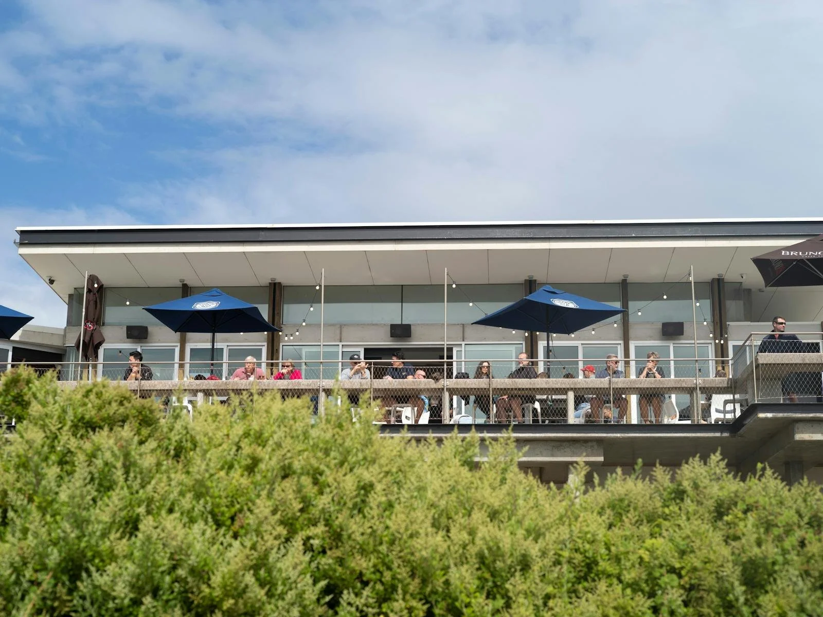 People sitting at tables on an outdoor balcony of a modern building, with umbrellas and string lights, under a partly cloudy sky.