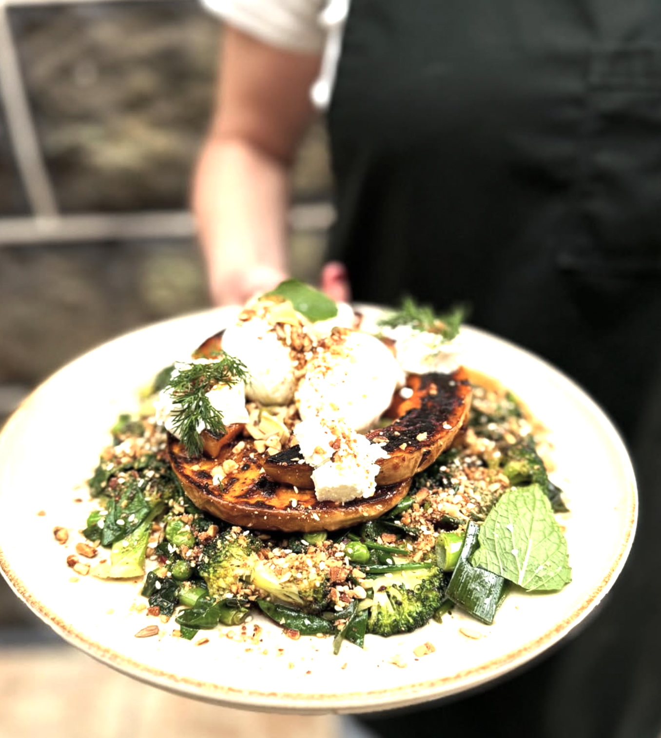 Close-up of a plate of grilled vegetables topped with herbs, nuts, and possibly goat cheese, held by a person wearing a black shirt.