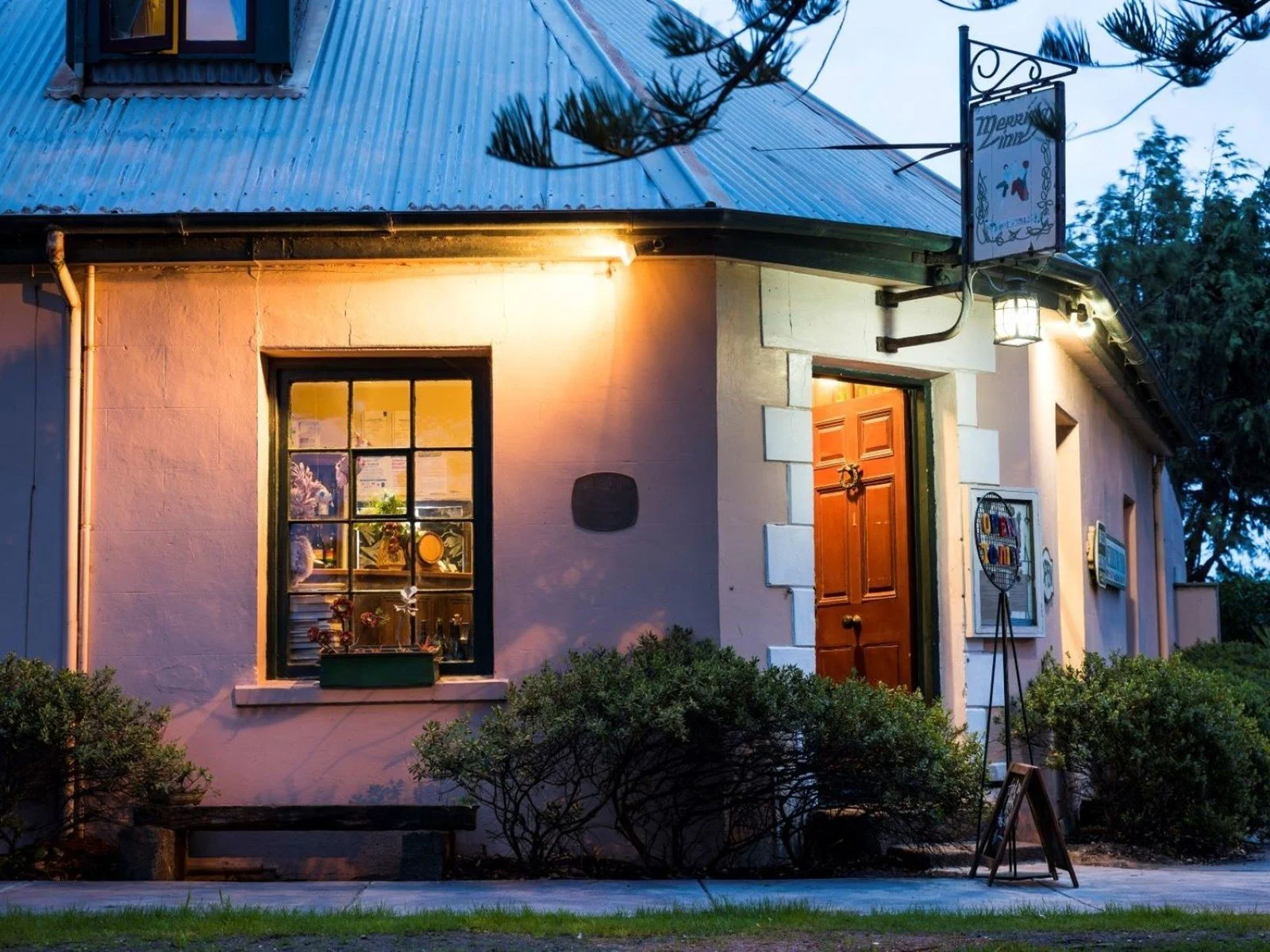 A cozy house with a lit wooden front door, a window with plants and decorations, and a sign hanging above. Outside are bushes and a sidewalk.
