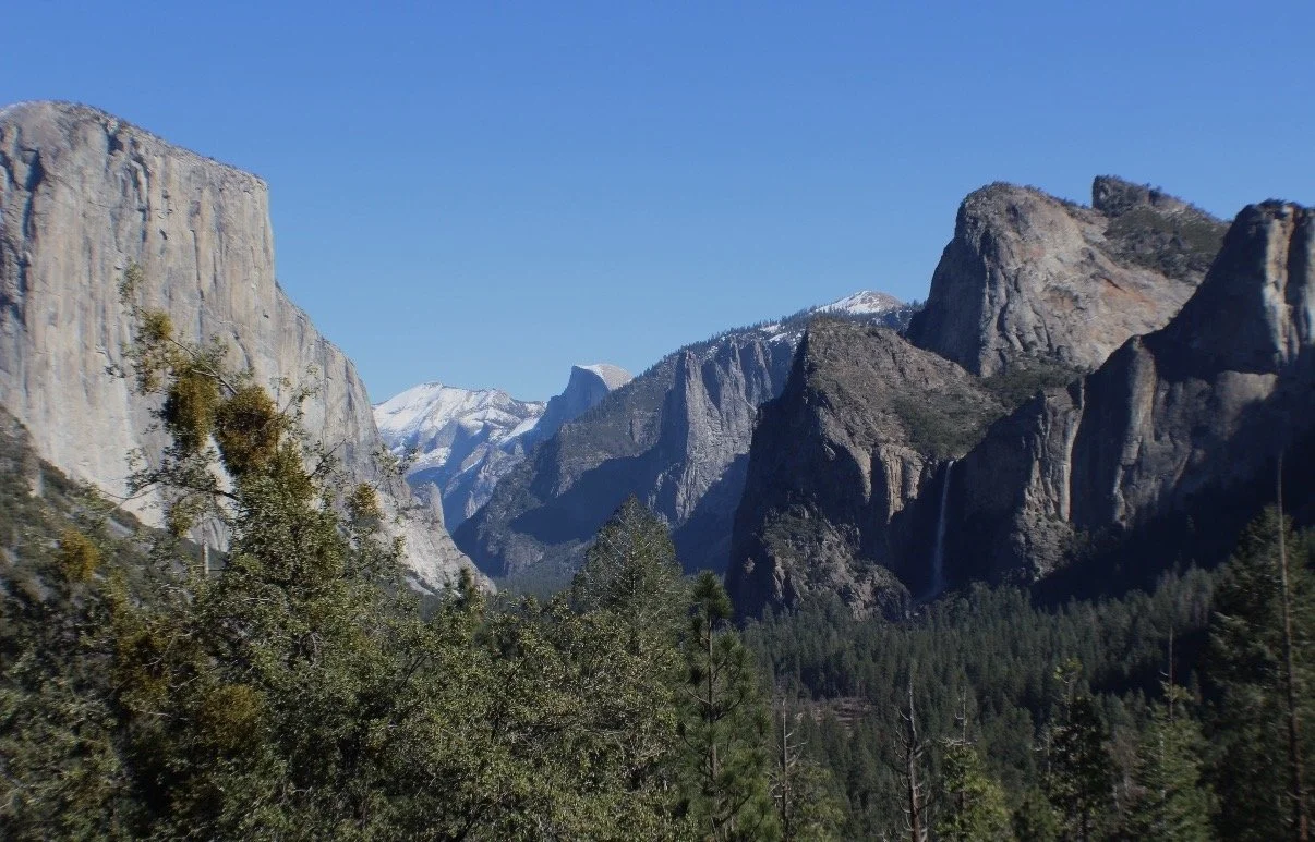 A panoramic view of a mountainous landscape with tall, rugged cliffs, snow-capped peaks, and dense green forests. The sky is clear and blue.