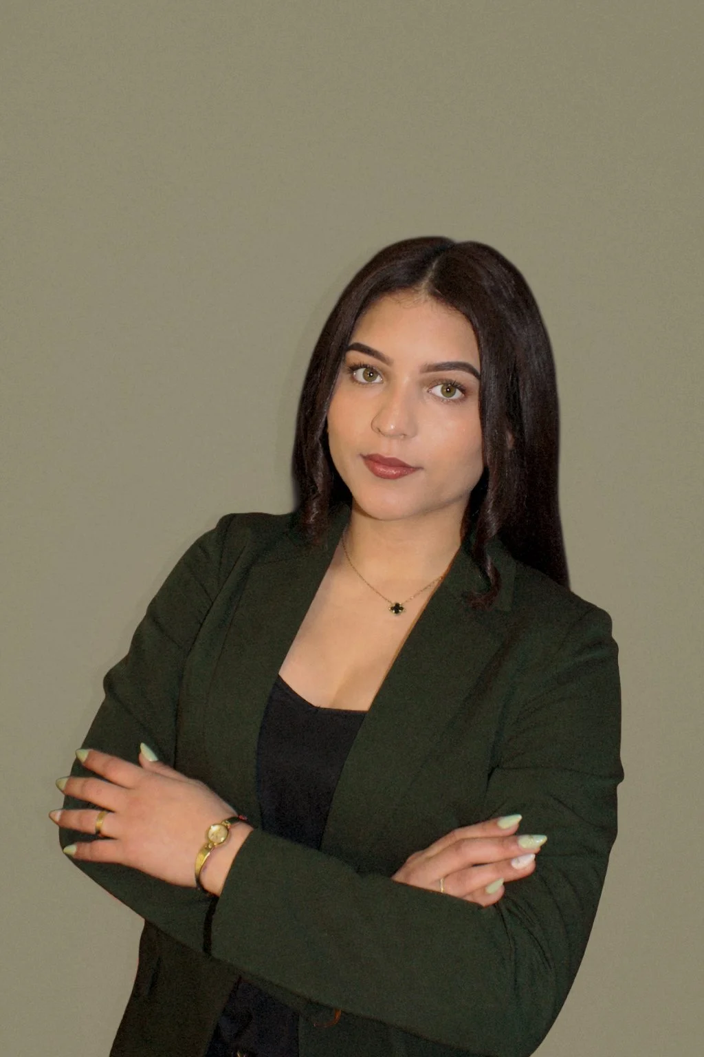 A woman with dark hair and green eyes wearing a black blazer, black top, and gold jewelry, standing against a plain gray background.
