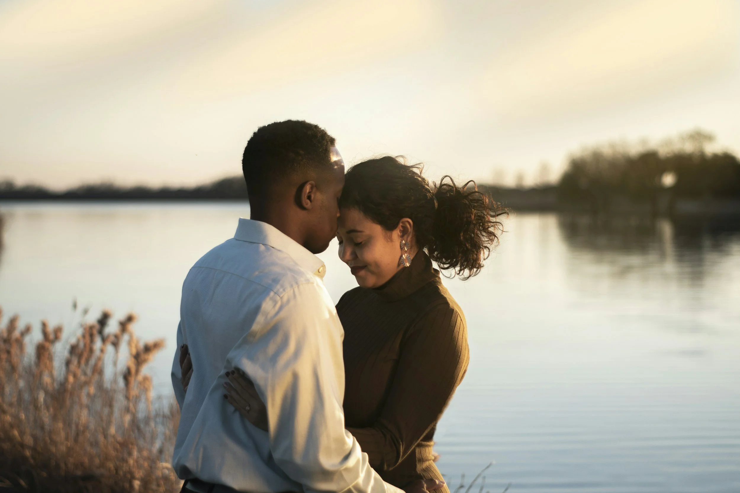 A couple standing close together with foreheads touching, near a body of water at sunset, embracing affectionately.