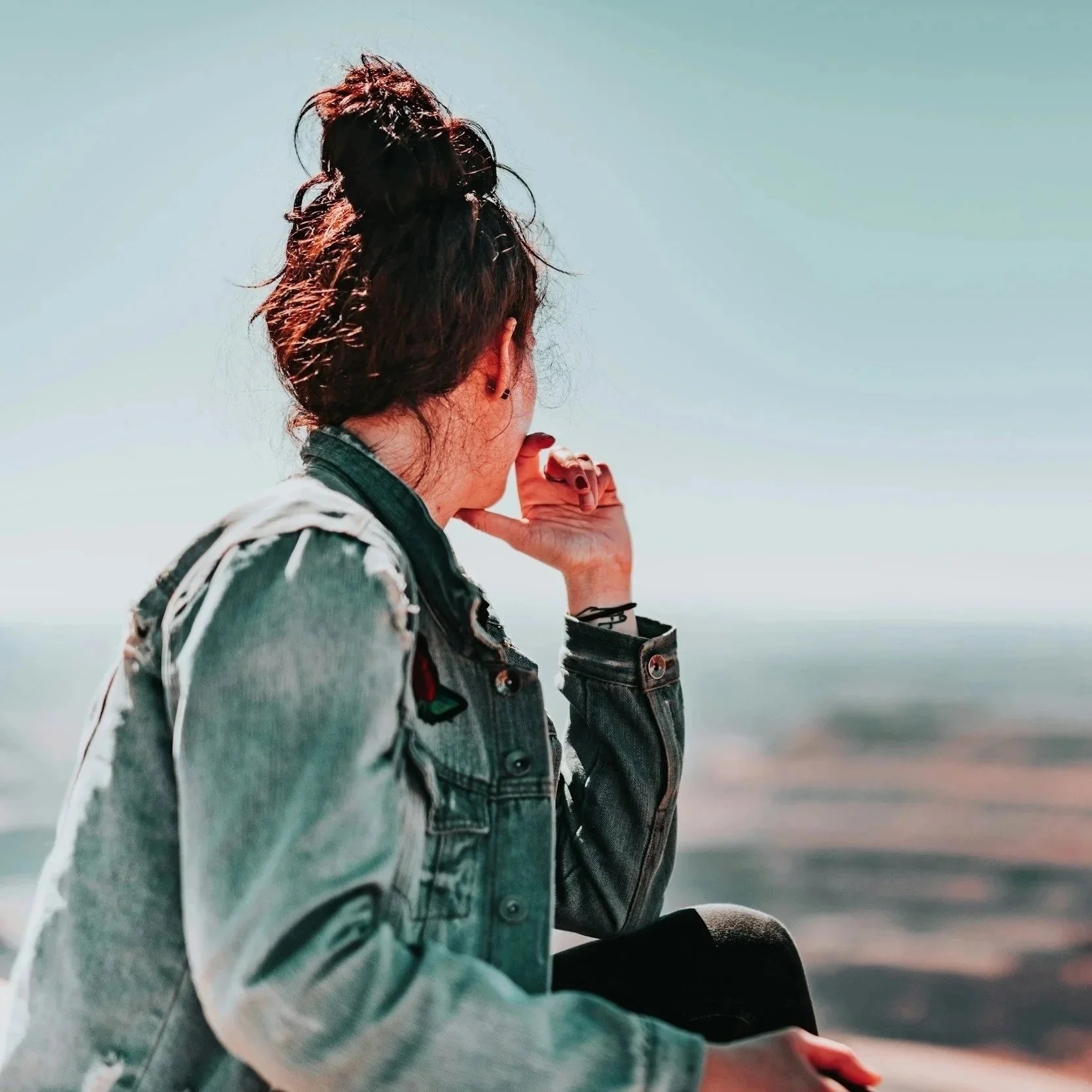 A woman with brown hair tied in a messy bun, wearing a denim jacket, sitting outdoors near water, looking into the distance with her hand resting on her face.