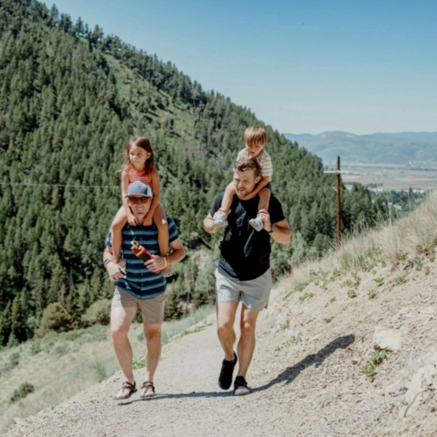 Couple hiking, one with a girl on his shoulders and another with a boy on his shoulders, walking along a dirt trail with mountains and trees in the background.