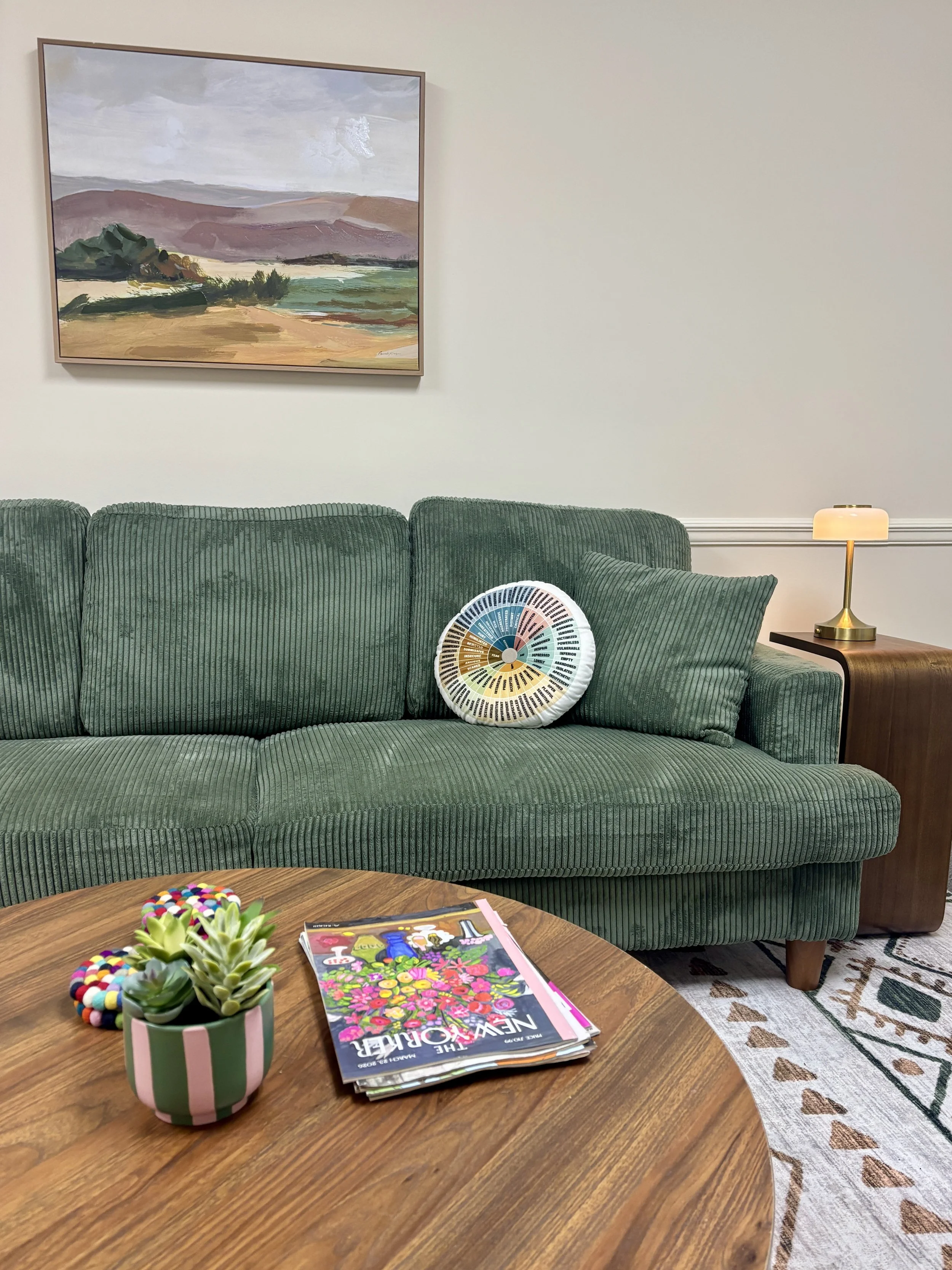 A living room with green velvet sofa, artwork of mountains and fields on the wall, small side table with a gold lamp, wooden coffee table with a potted succulent and colorful magazine.