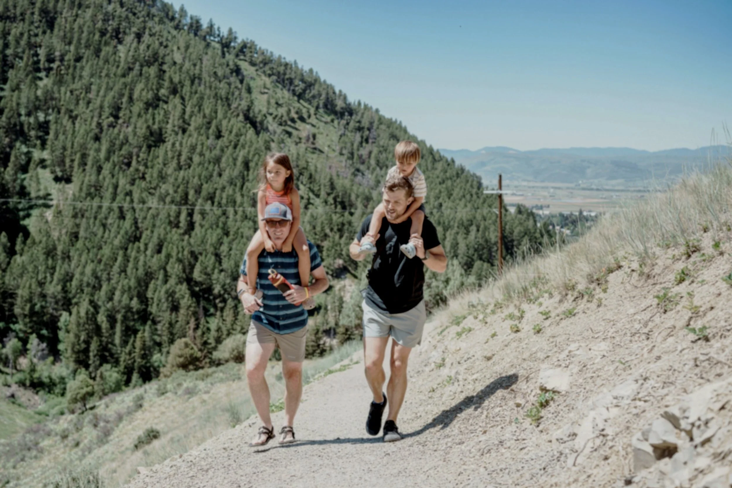 Couple hiking on a dirt trail in a mountainous area with two children, one girl sitting on a man's shoulders and a boy sitting on another man's shoulders, alongside a backdrop of dense green forest and distant mountains under a clear blue sky.