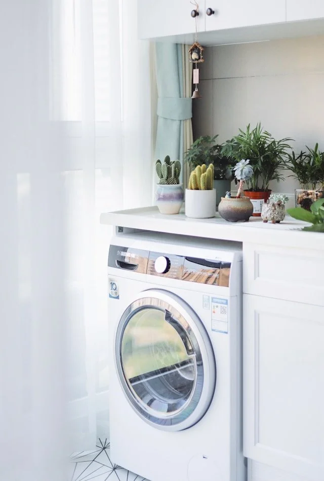 Front view of a white washing machine beneath a white countertop with potted plants on top, set in a bright room with curtains and a window.