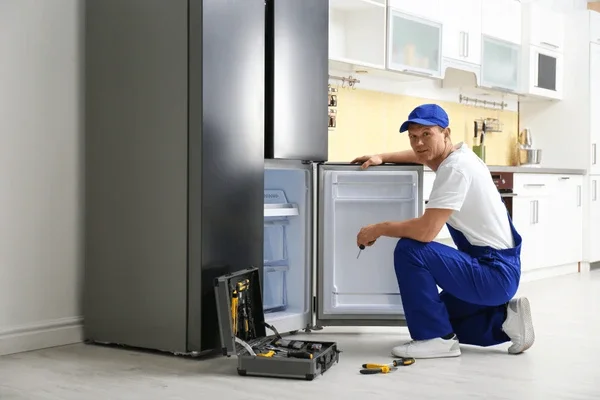 A technician repairing a refrigerator in a modern kitchen, kneeling next to the open fridge door with tools on the floor.
