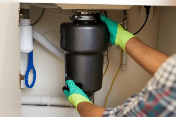 A person wearing yellow and green gloves installing or repairing a garbage disposal unit under a kitchen sink.