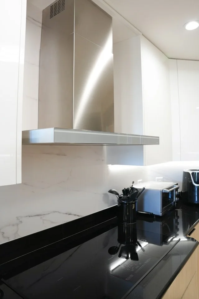 Modern kitchen counter with black countertop, stainless steel range hood, white cabinetry, and small appliances including a toaster, pitcher, and utensil holder.