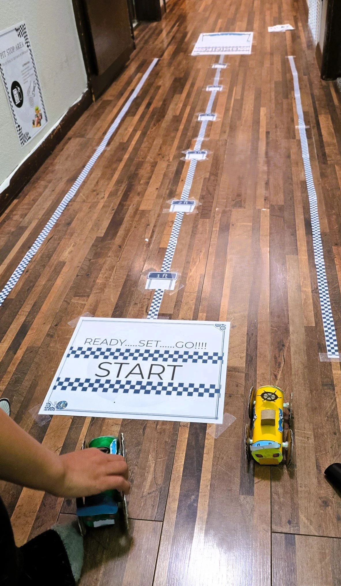 A floor with checkered tape lines and signs for a racing activity, including a large sign that says 'READY...SET...GO!!!! START.' Two remote control toy cars are positioned at the starting line, with a child’s hand nearby.
