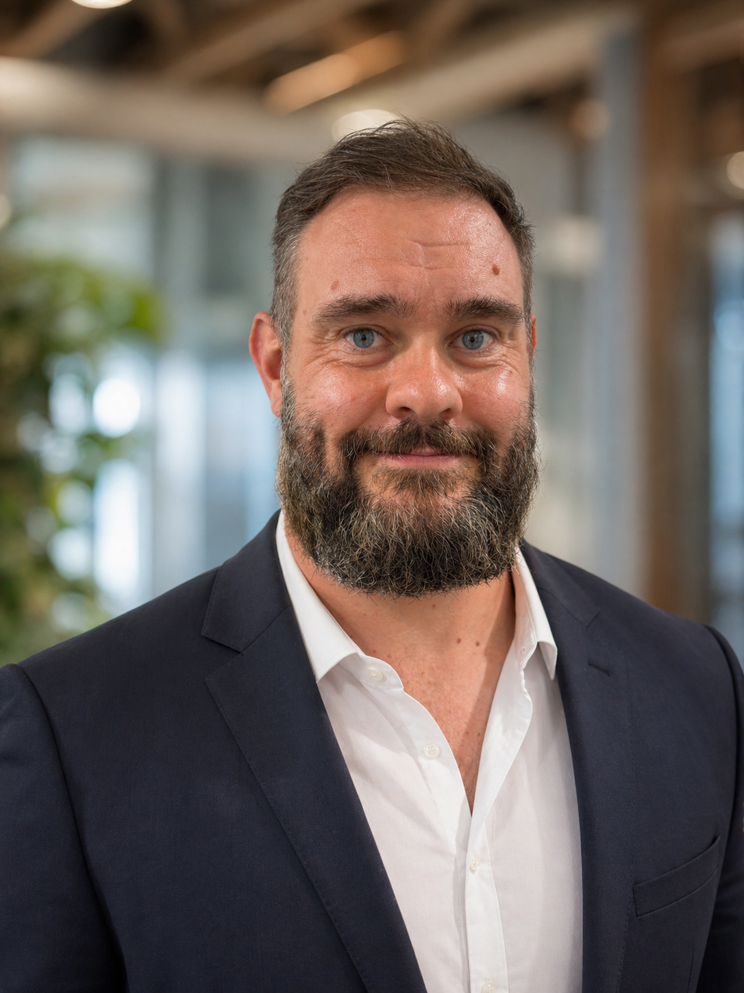 Headshot of a man with a beard in a navy blazer and white shirt, smiling indoors with blurred background.