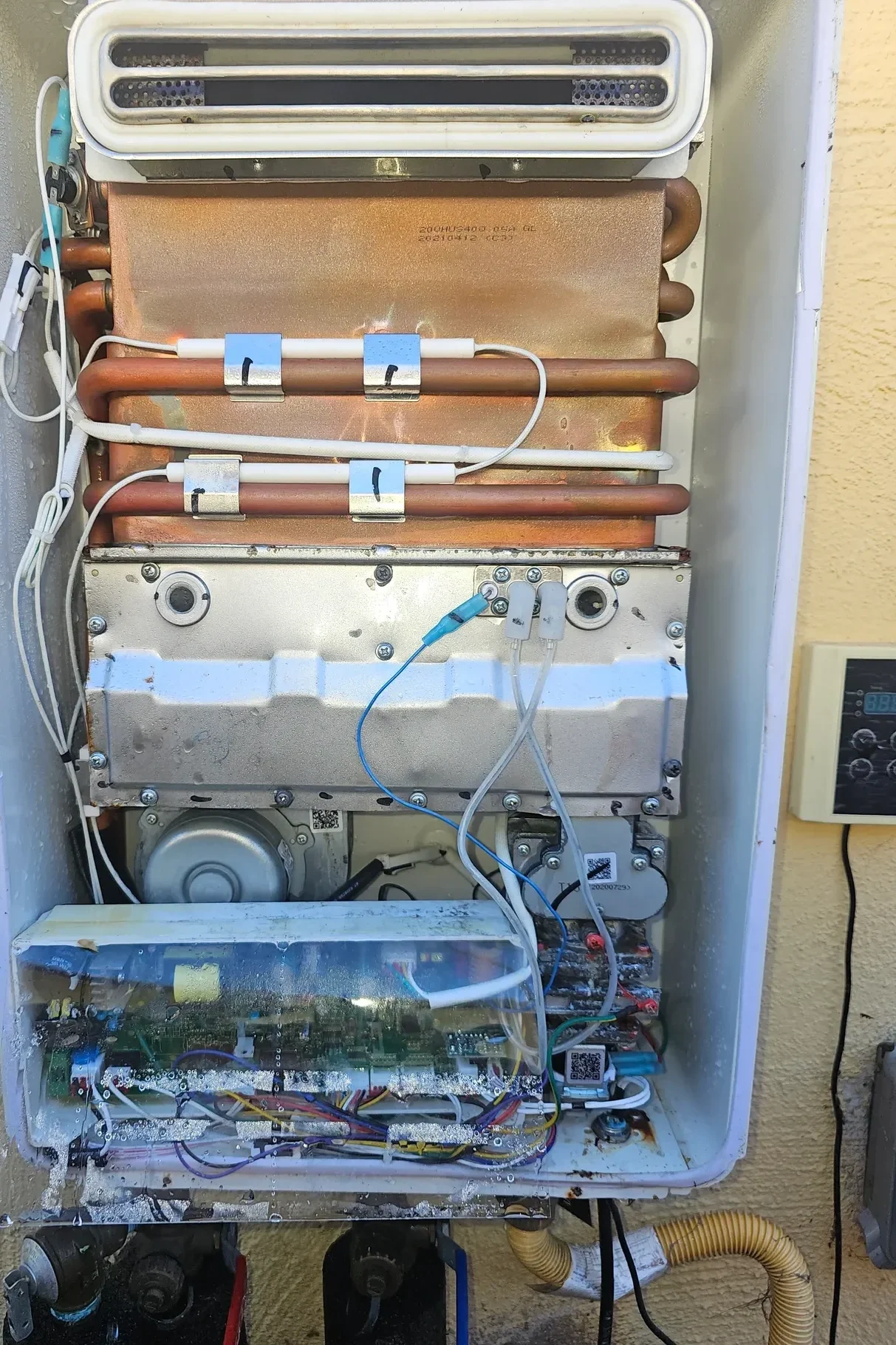 Open outdoor air conditioning unit with exposed inner components, wires, and copper coils inside a metal casing, mounted on a wall.