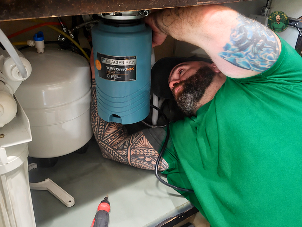 Man with a tattooed arm, beard, and green shirt working under kitchen sink, repairing or installing plumbing, with tools nearby.