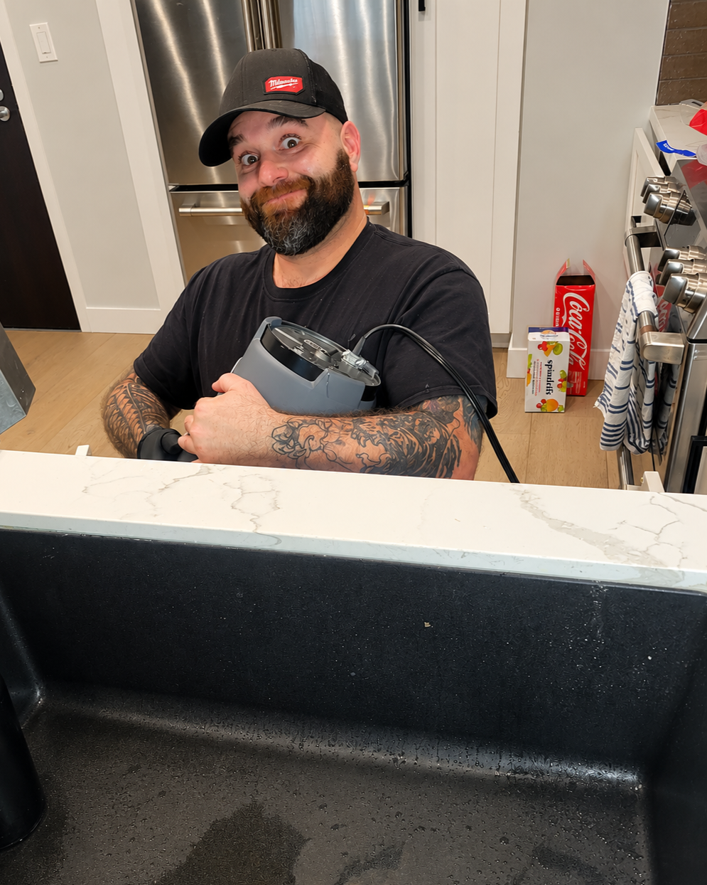A man with tattoos on his arms holding a kitchen appliance in a modern kitchen, smiling at the camera, wearing a black T-shirt and a black baseball cap.