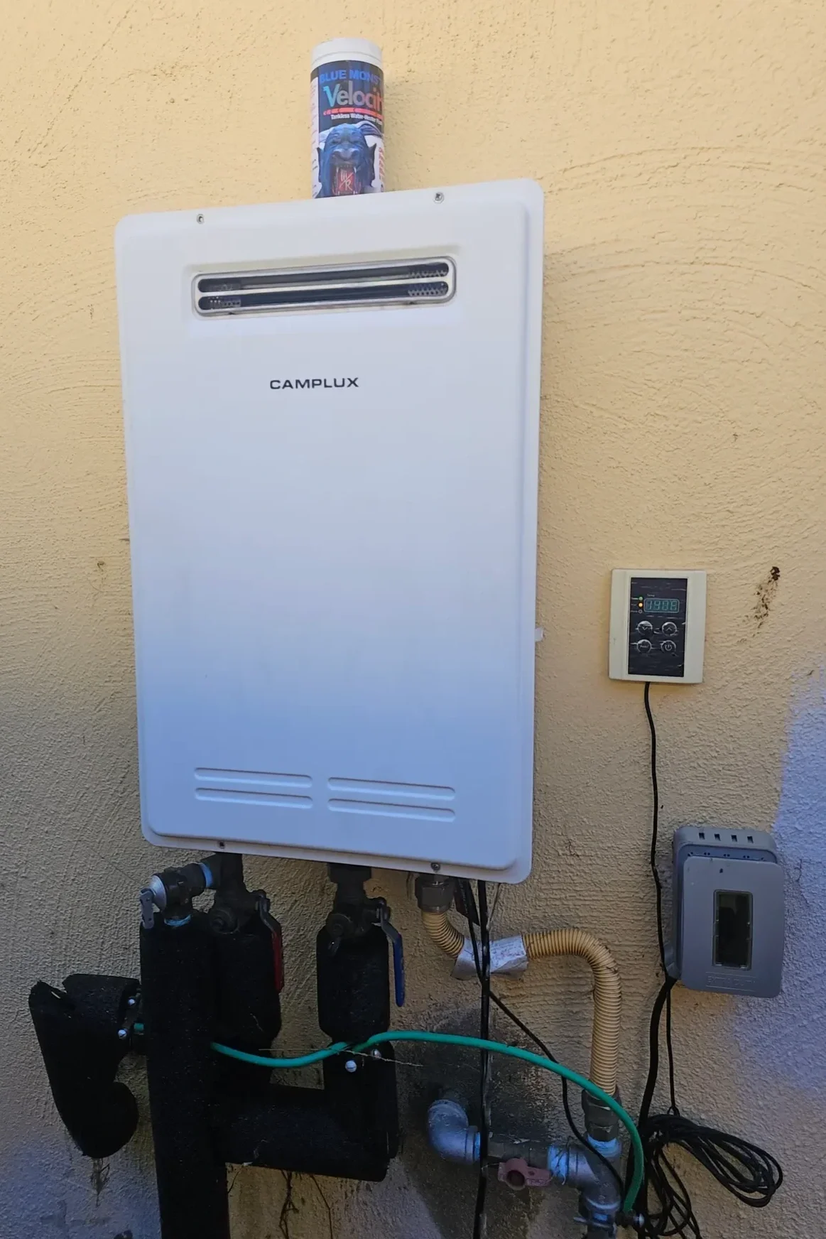 An outdoor HVAC system with a white Camplux tankless water heater, a thermostat, and various pipes and controls mounted on a yellow wall. A container of Velociraptor toothpaste is placed on top of the heater.