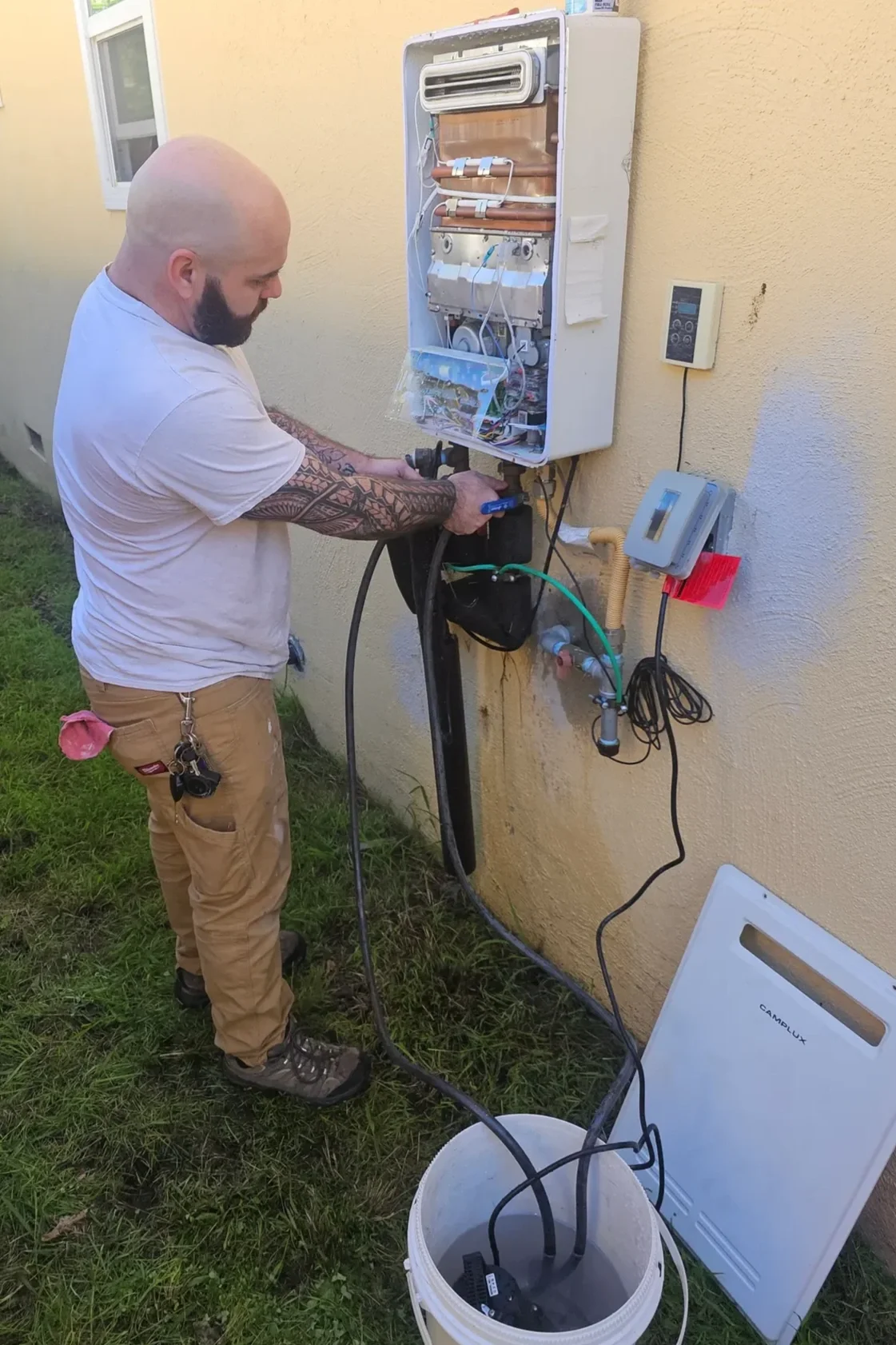 A man with tattoos on his arm working on an outdoor air conditioning or refrigeration unit, using tools. The unit's cover is removed, exposing internal components. The scene shows various electrical and plumbing connections on a beige wall, with a digital control panel nearby and a white appliance on the ground.