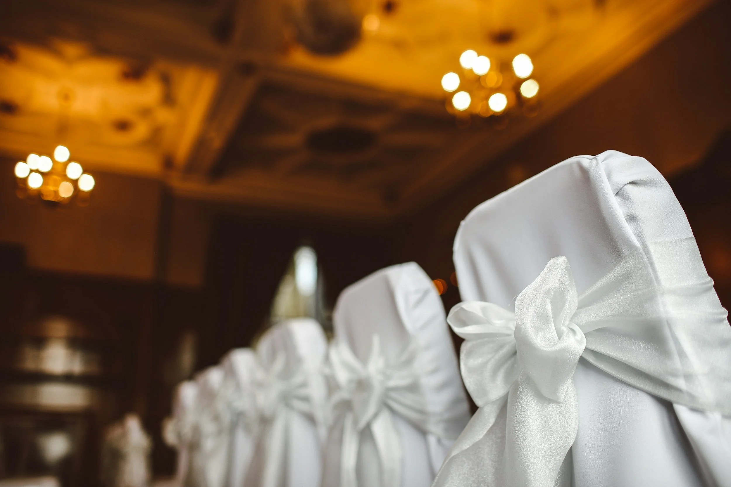 Rows of white chair covers in a banquet hall with a decorated ceiling and chandelier lighting