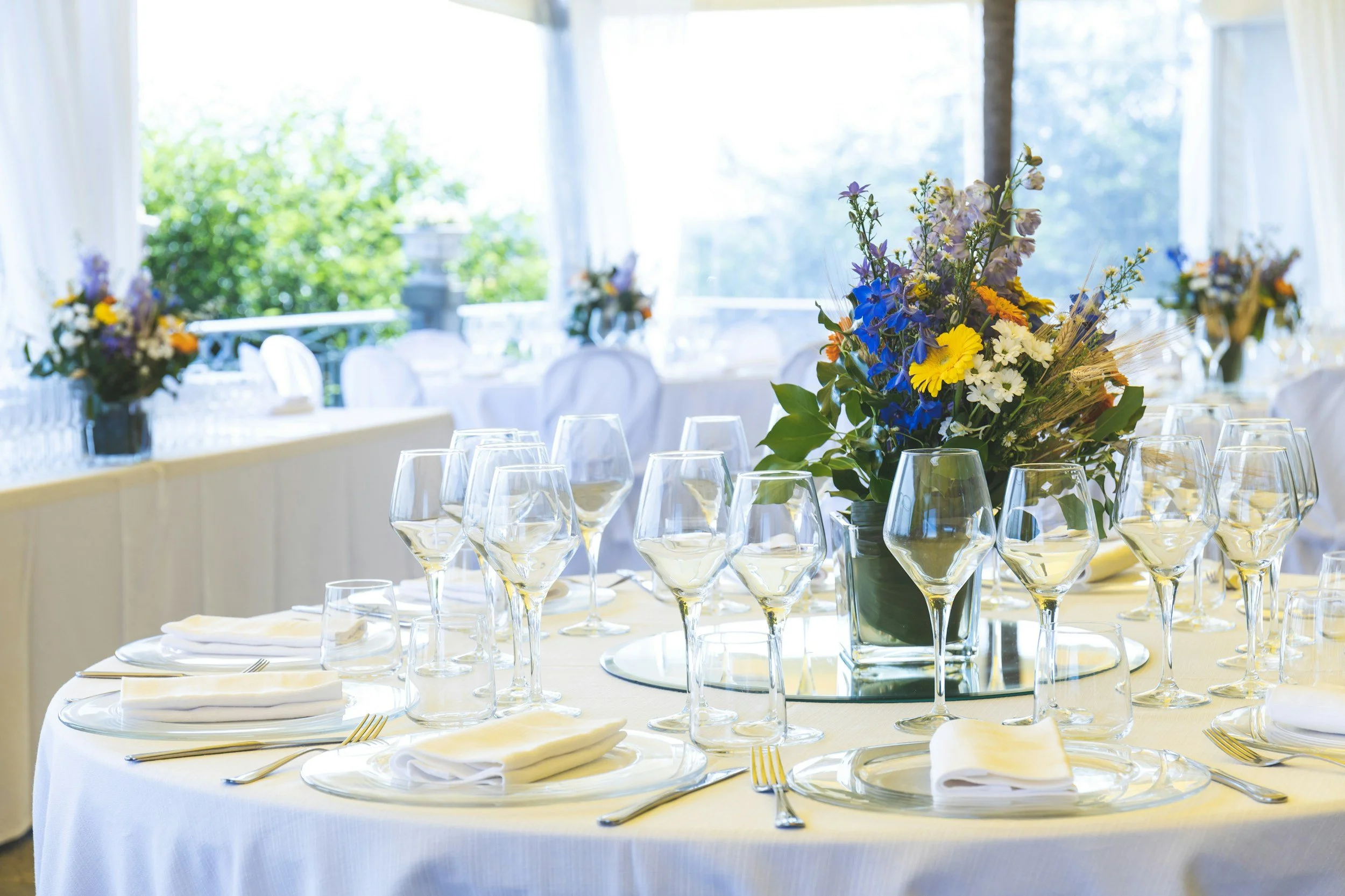 A banquet table set with white tablecloths, neatly folded white napkins, wine glasses, and gold utensils, with a central floral arrangement of mixed colorful flowers in a green vase, outside view with trees through large windows.