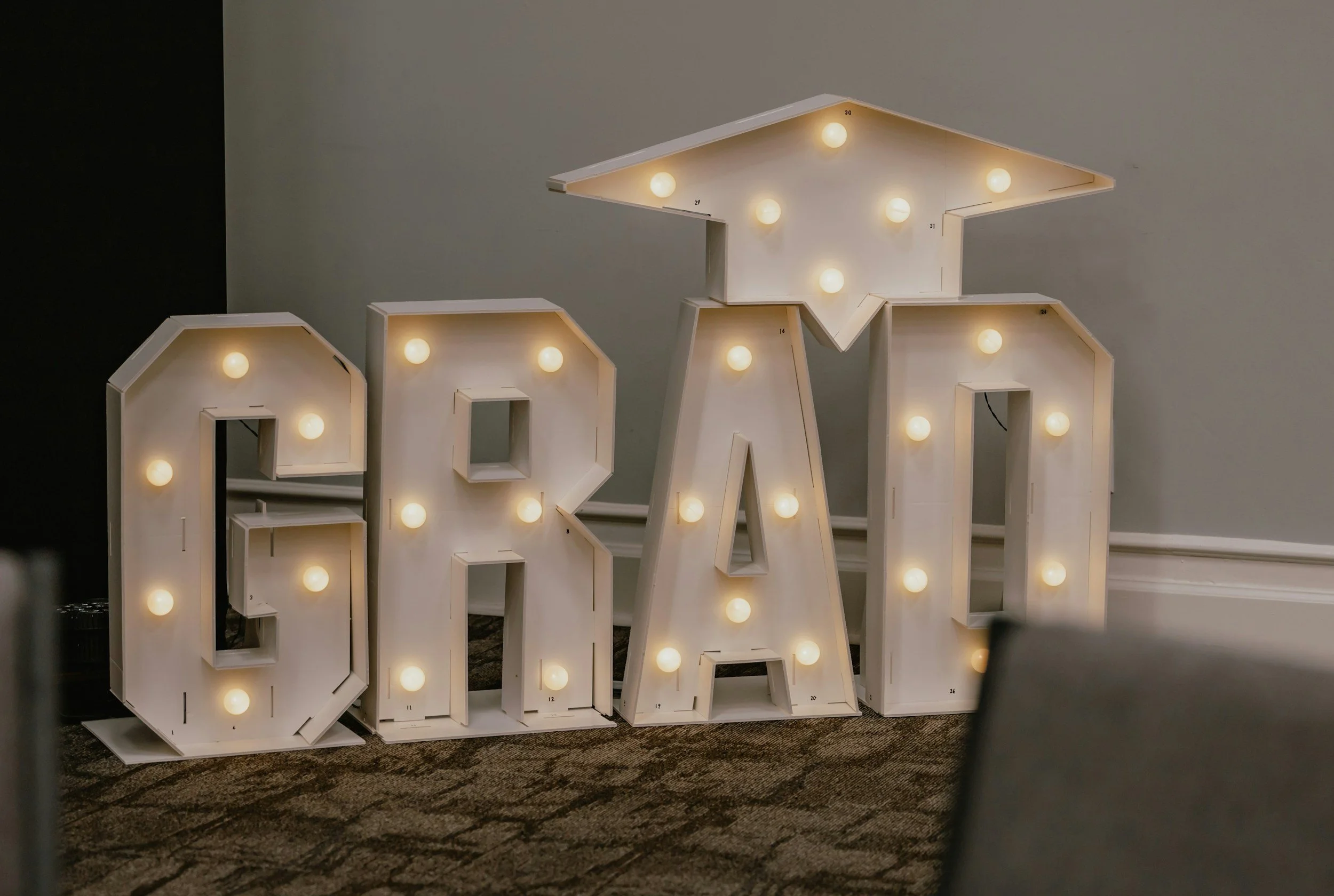 Decorative 'LOVE' sign made of white wooden letters with warm light bulbs on each letter, placed on a carpeted floor against a plain wall.