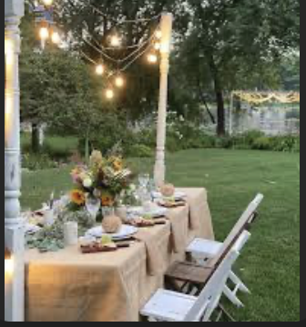 Outdoor dining table set with a beige tablecloth, floral centerpiece, candles, plates, and silverware, under string lights in a garden