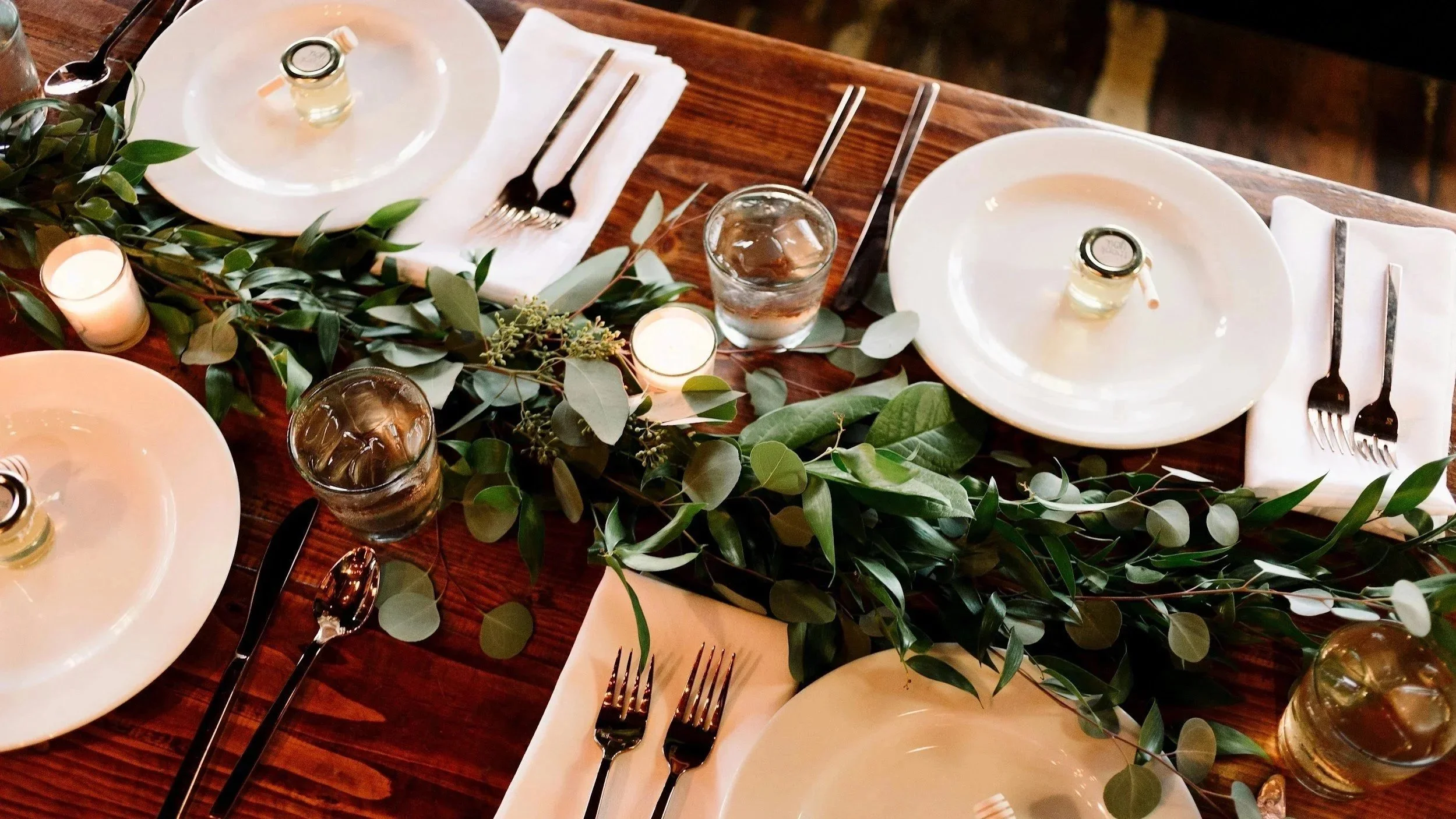 A decorated dining table with white plates, silverware, glasses of water with ice, candles, and greenery garland for a formal event.