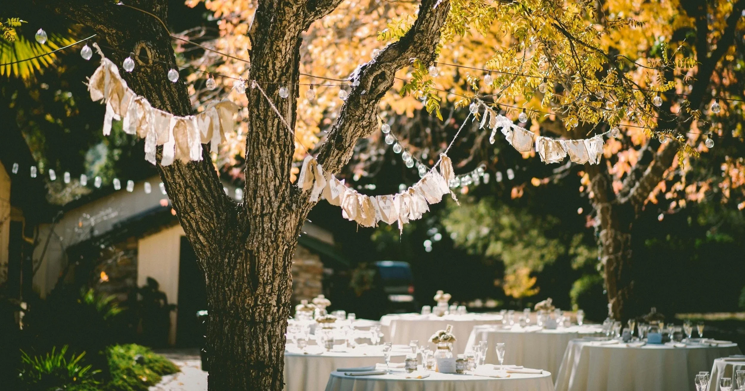 Outdoor garden party setup with tables decorated with white tablecloths, surrounded by trees with autumn leaves, string lights, and garlands hanging from the trees.